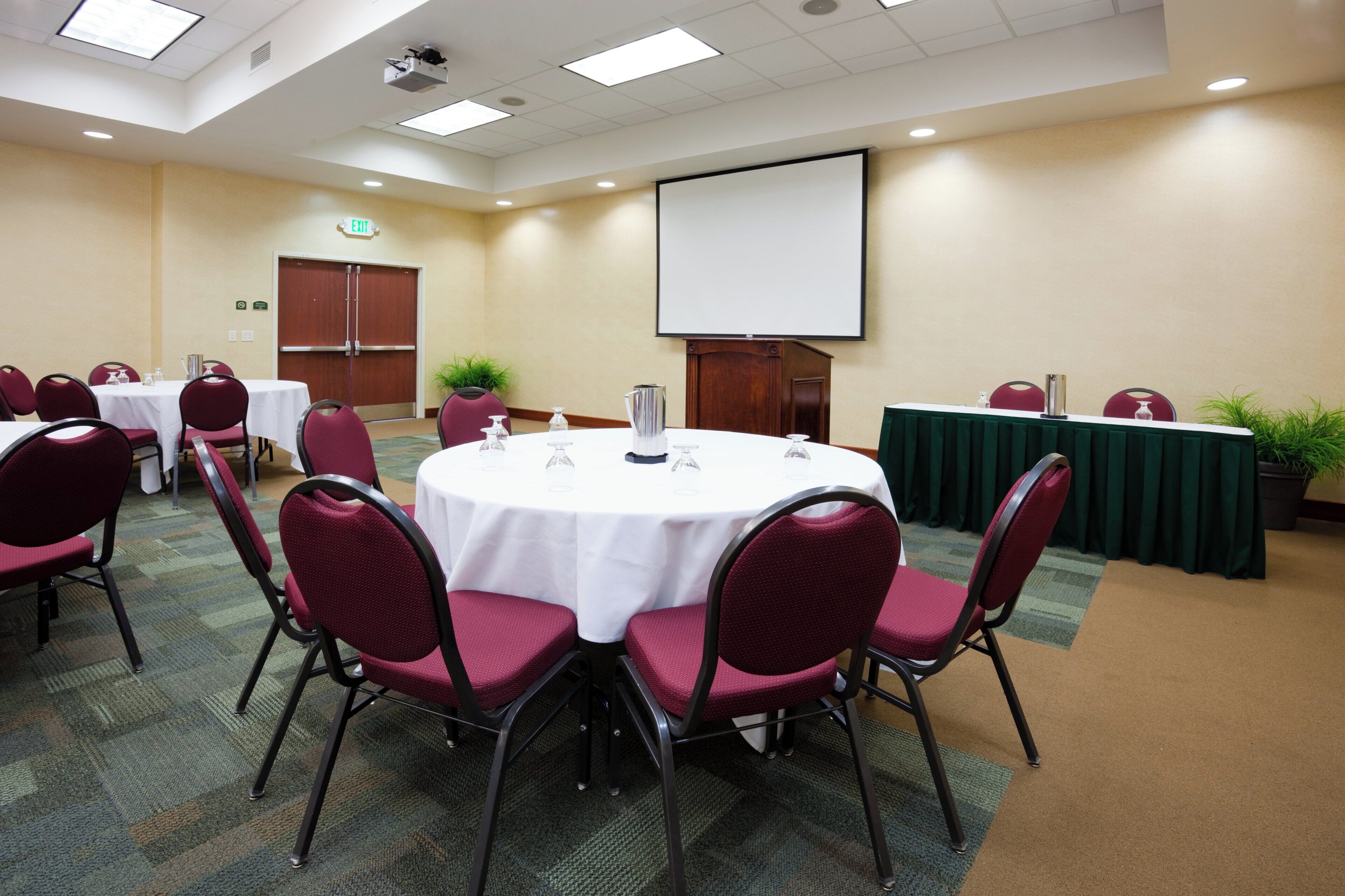 A conference room with round tables, chairs, a projector screen, and an exit door.