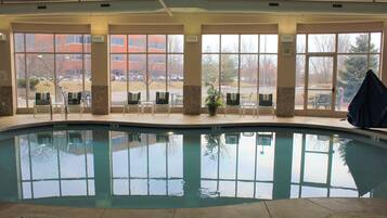 Una piscina interior con paredes de vidrio transparente, suelo de baldosas y vistas a un estacionamiento y edificios.