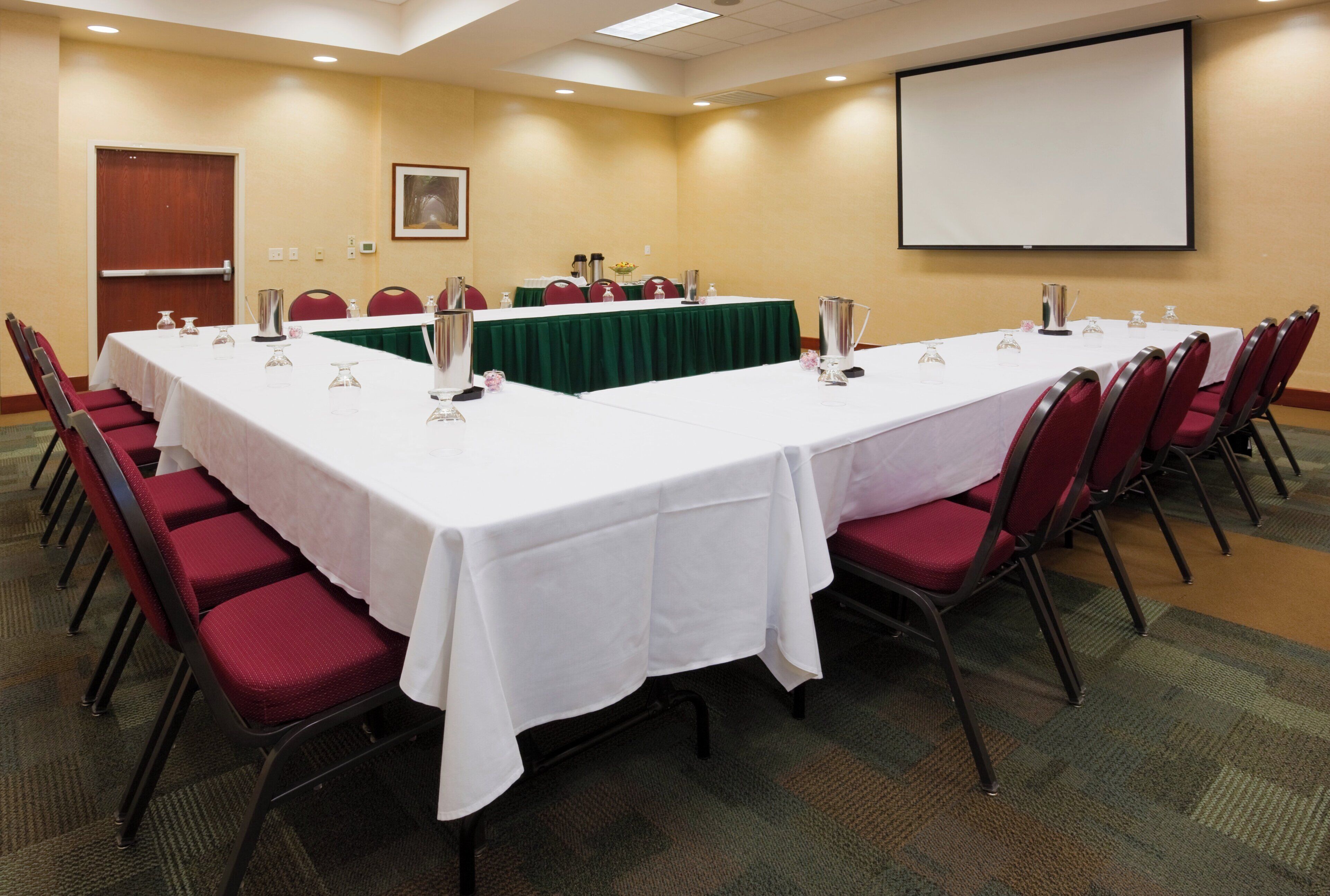 A conference room with a long table, red chairs, and a projector screen.