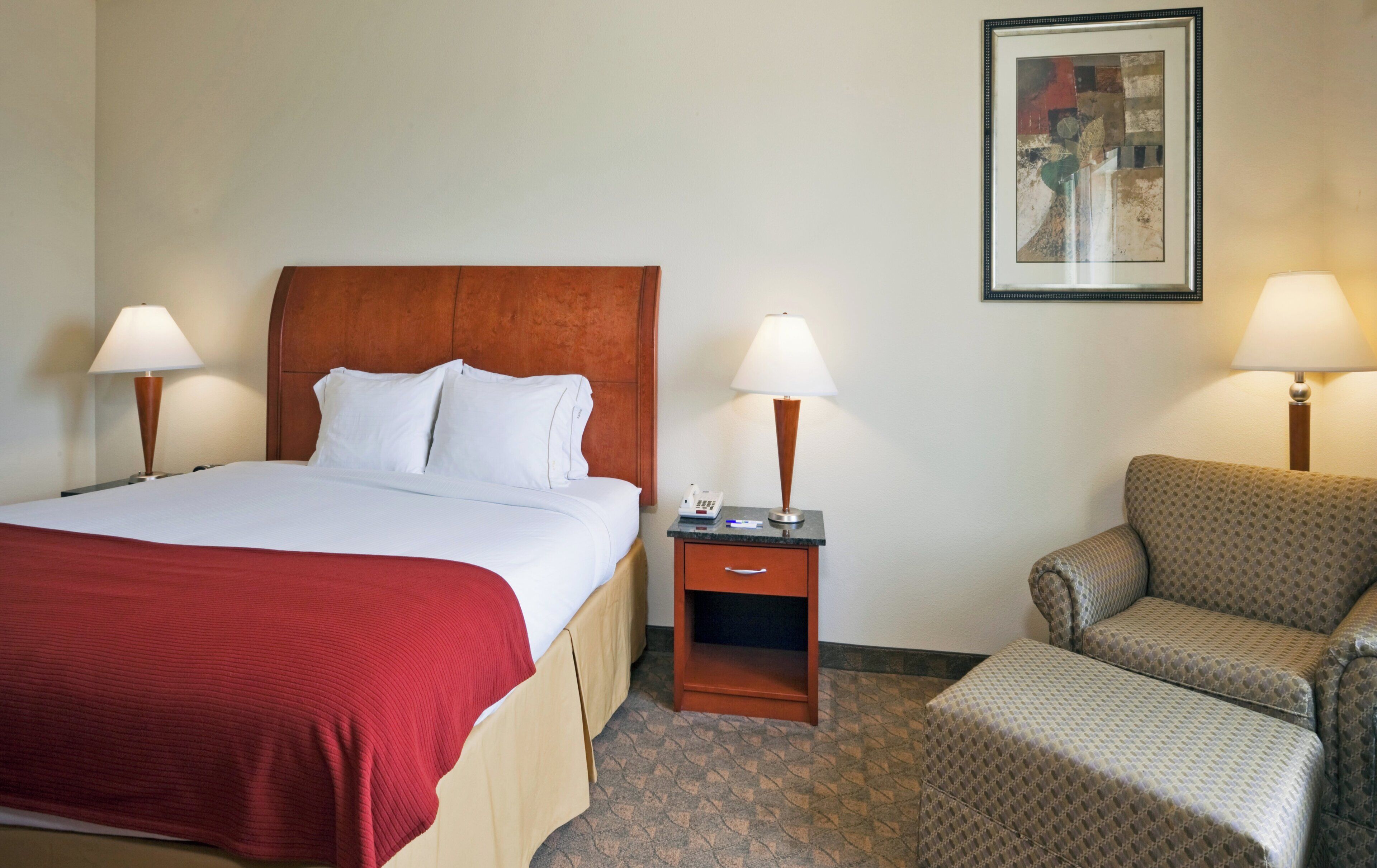 A hotel room with a bed, two bedside lamps, a nightstand, a chair, and a painting on the wall.
