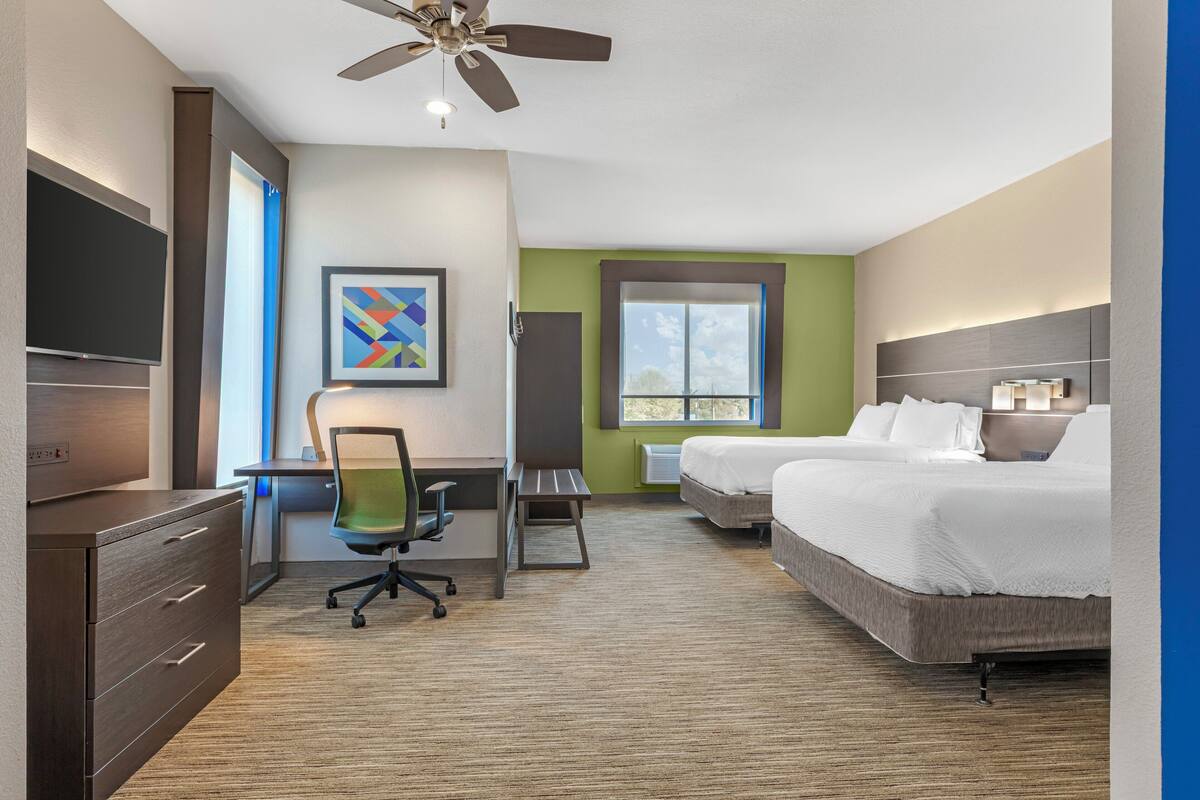 A hotel room with a desk, chair, two beds, a ceiling fan, and a TV.