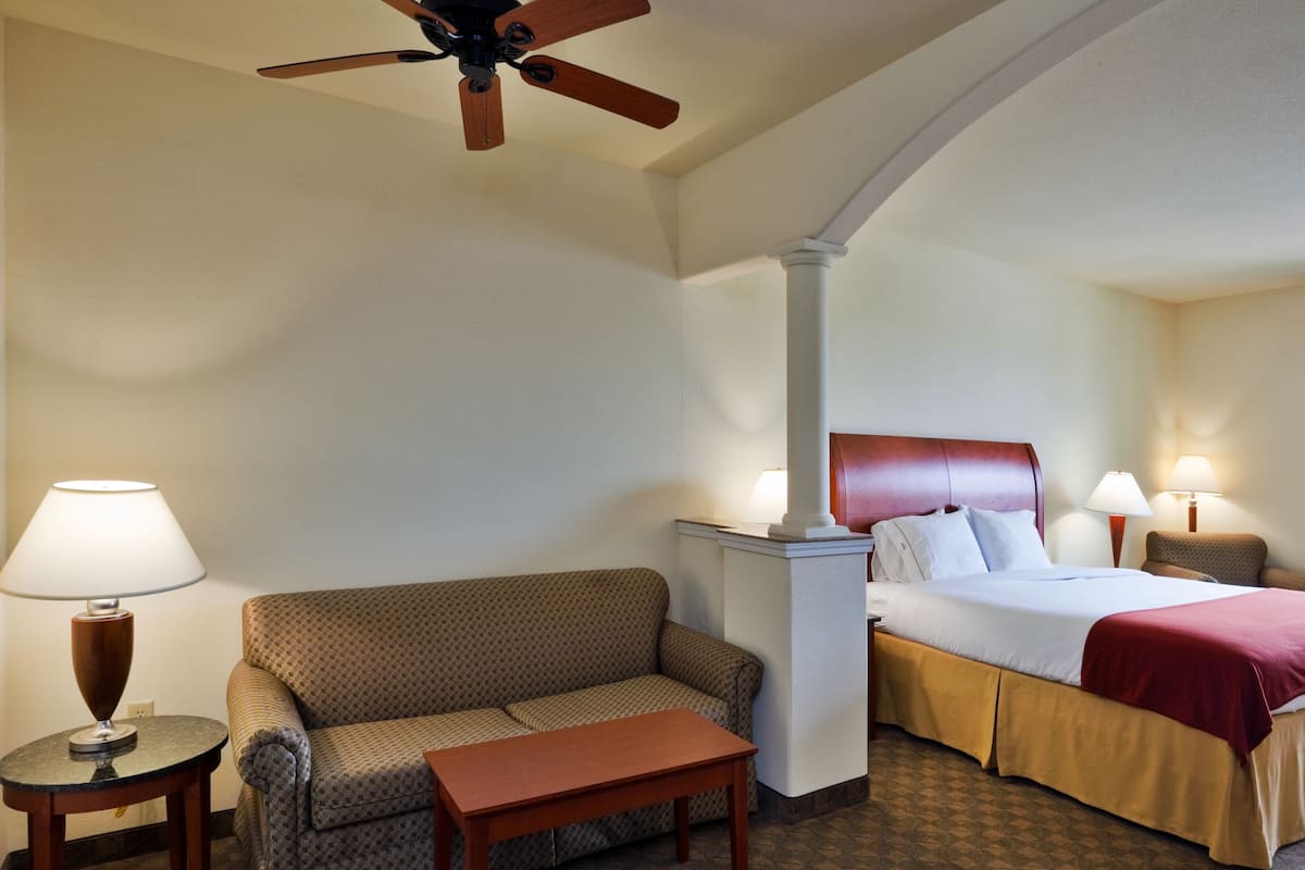 A hotel room with a bed, a sofa, a small table, a lamp, a ceiling fan, and a nightstand.