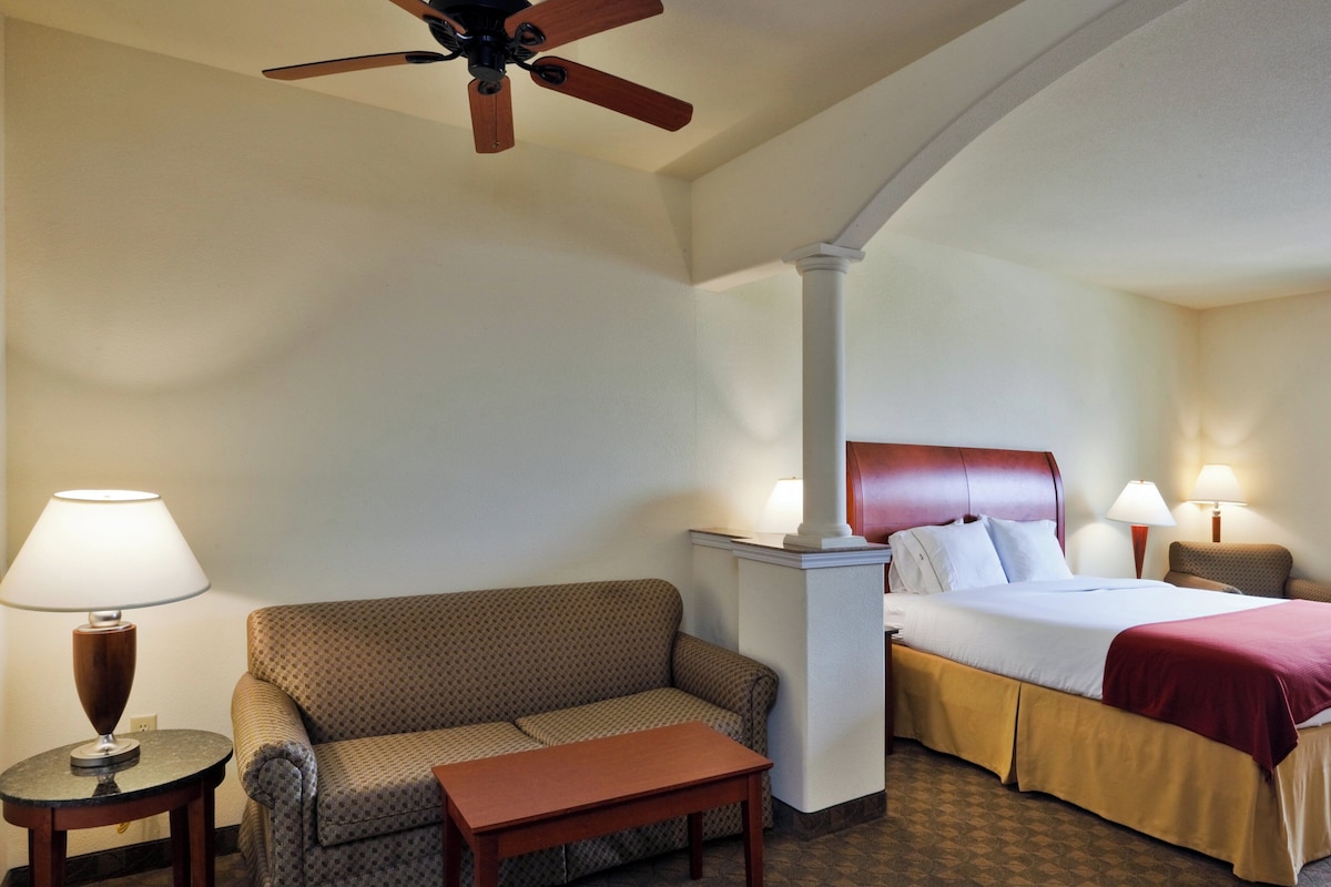 A hotel room with a bed, a sofa, a small table, a lamp, and a ceiling fan.