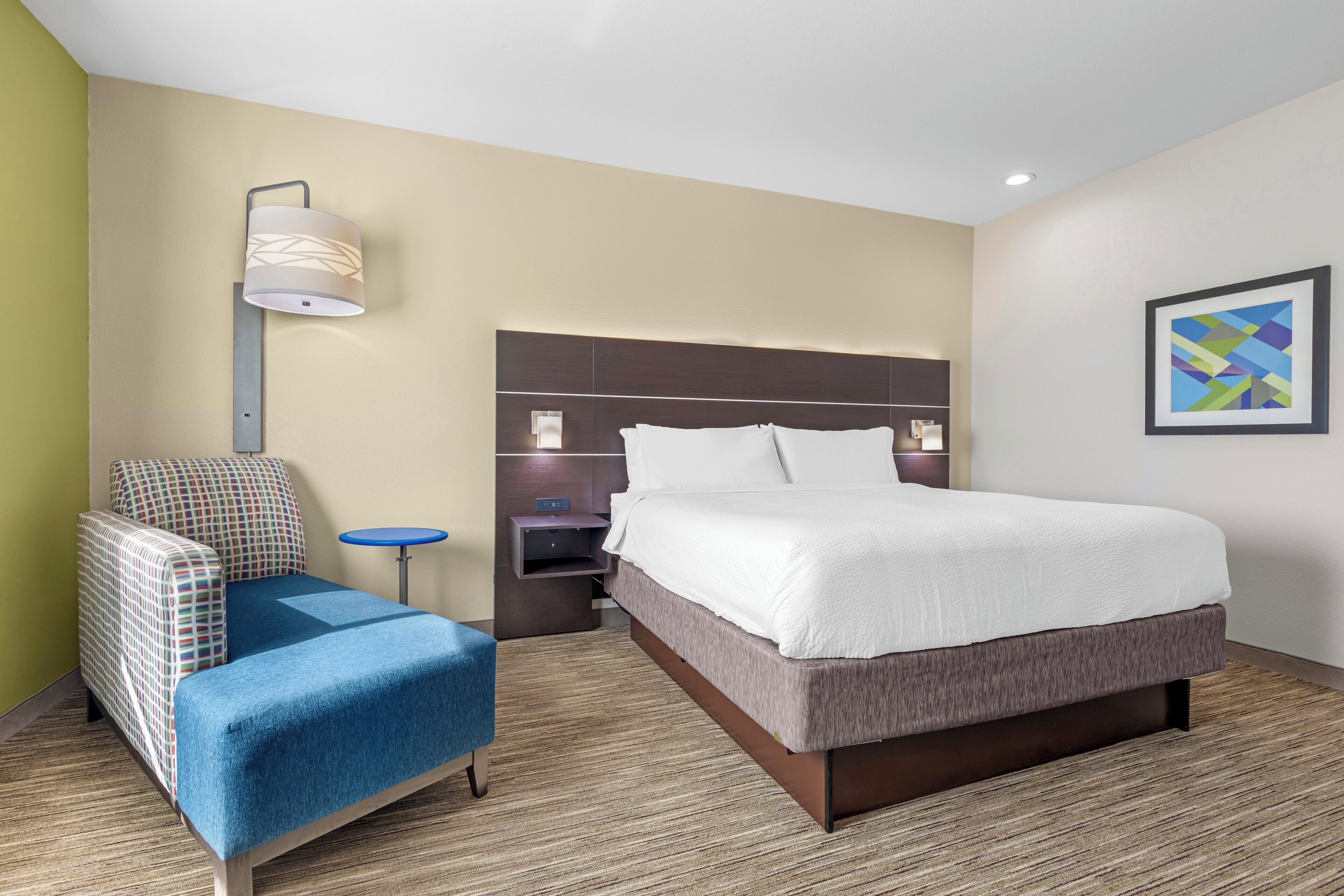 A hotel room with a bed, a blue armchair, a small table, a wall-mounted lamp, and a framed picture.