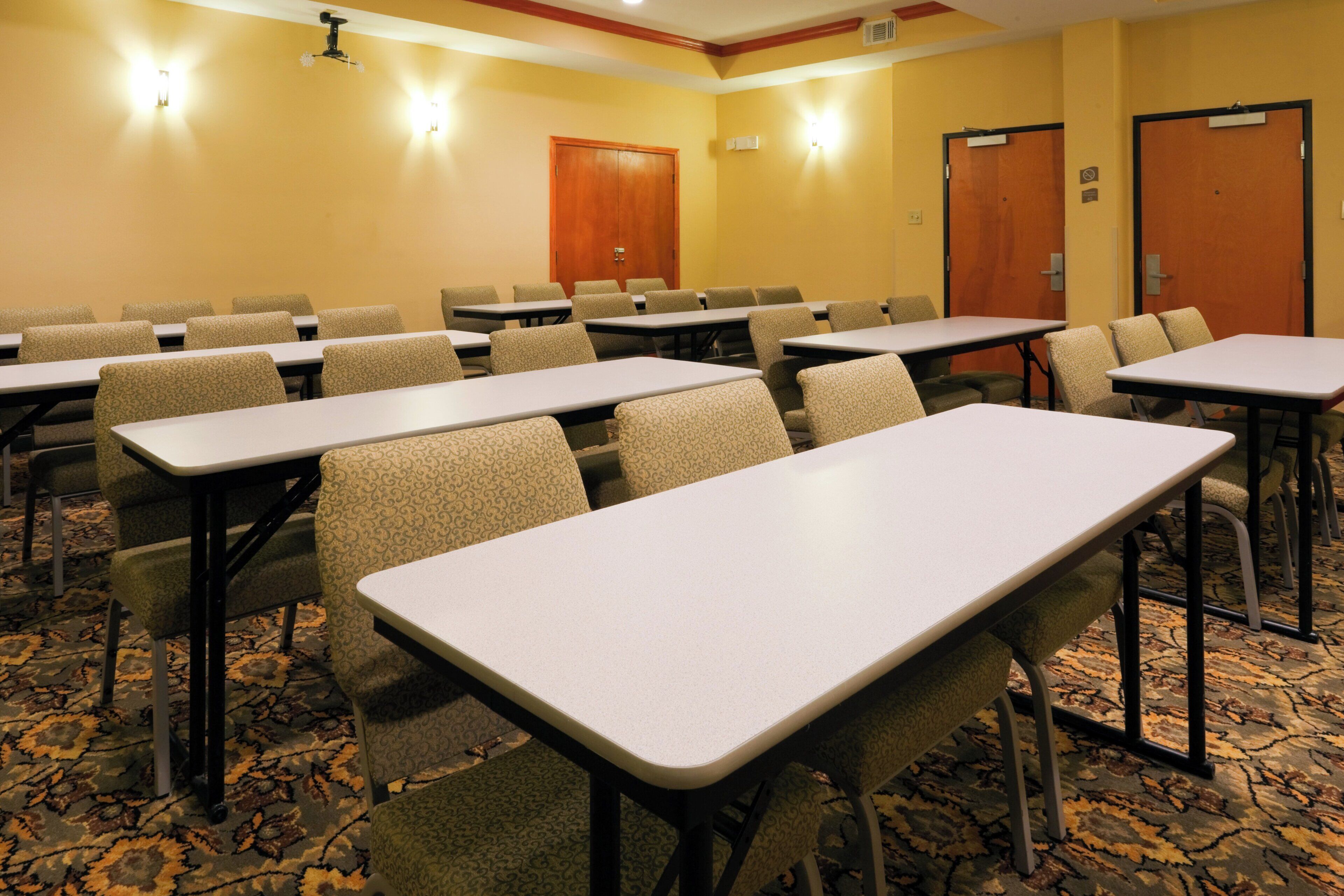 A conference room with multiple tables and chairs, carpeted flooring, and a wooden door in the background.