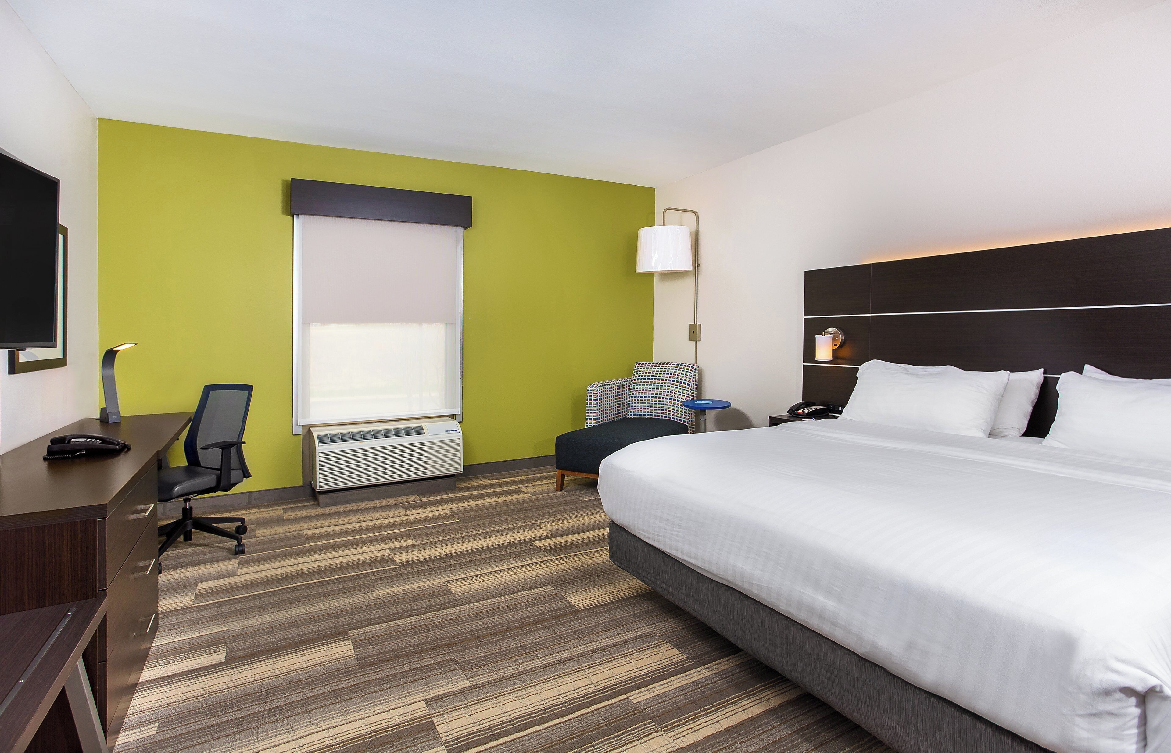 A hotel room with a large bed, a desk with a chair, a standing lamp, and a window with blinds.