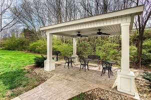 Un espacio cubierto con patio, con una mesa y sillas, rodeado de un jardín y árboles.