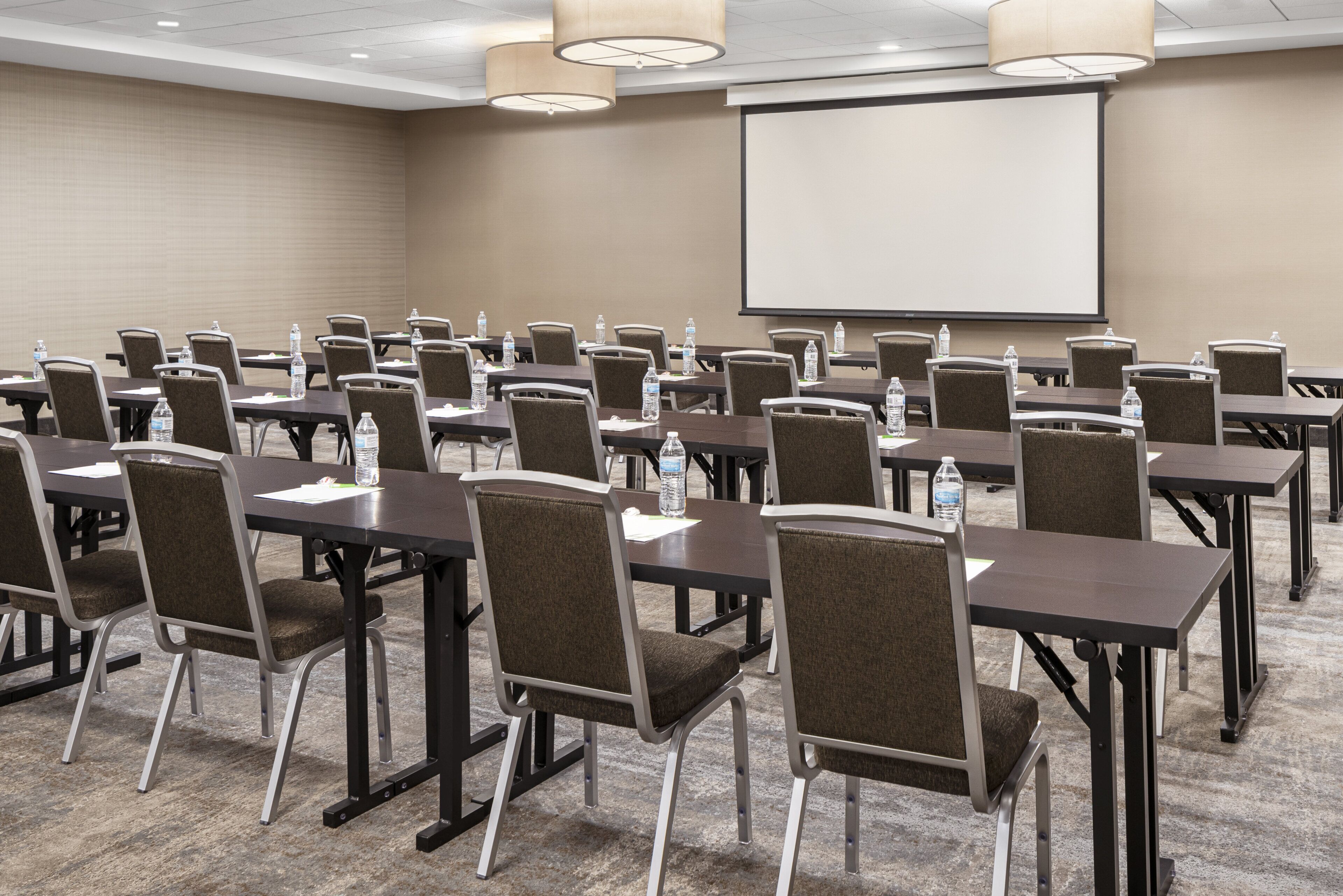 Un salón de conferencias con varias mesas y sillas, equipado con botellas de agua y documentos, y una pantalla de proyector.