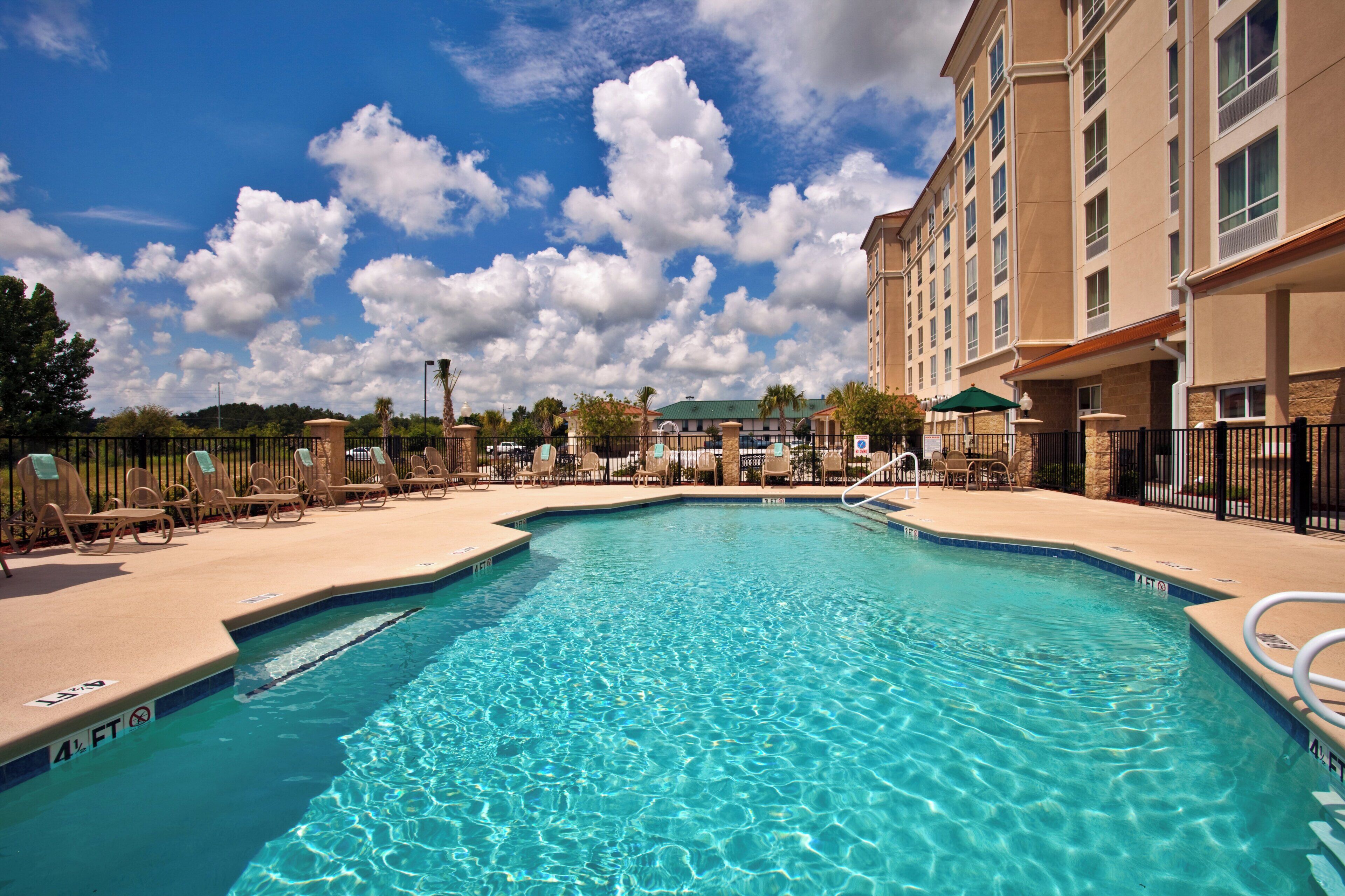 A clear blue pool with lounge chairs and umbrellas, surrounded by a hotel building.