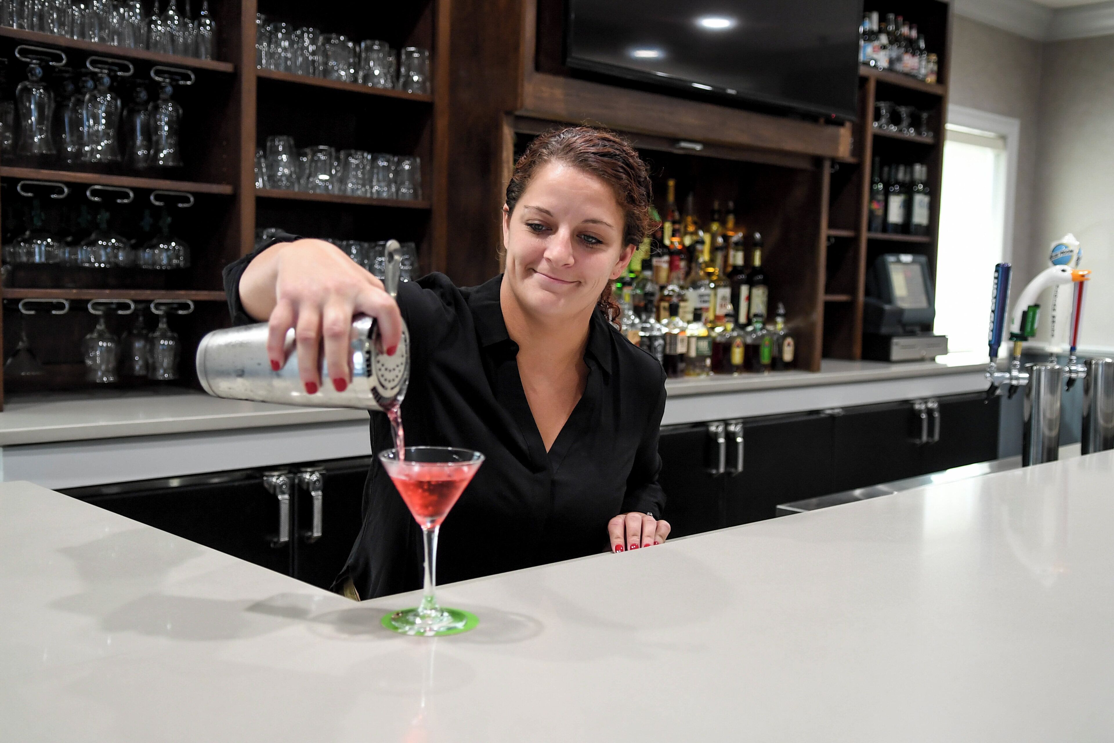 A bartender pouring a drink into a martini glass at a bar with shelves of bottles and glasses in the background.