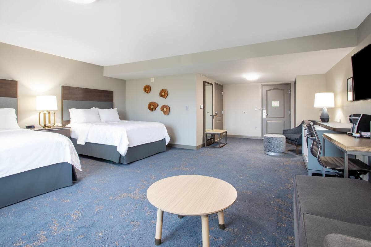 A hotel room with two beds, a round table, a desk with a chair, a TV, and a door.