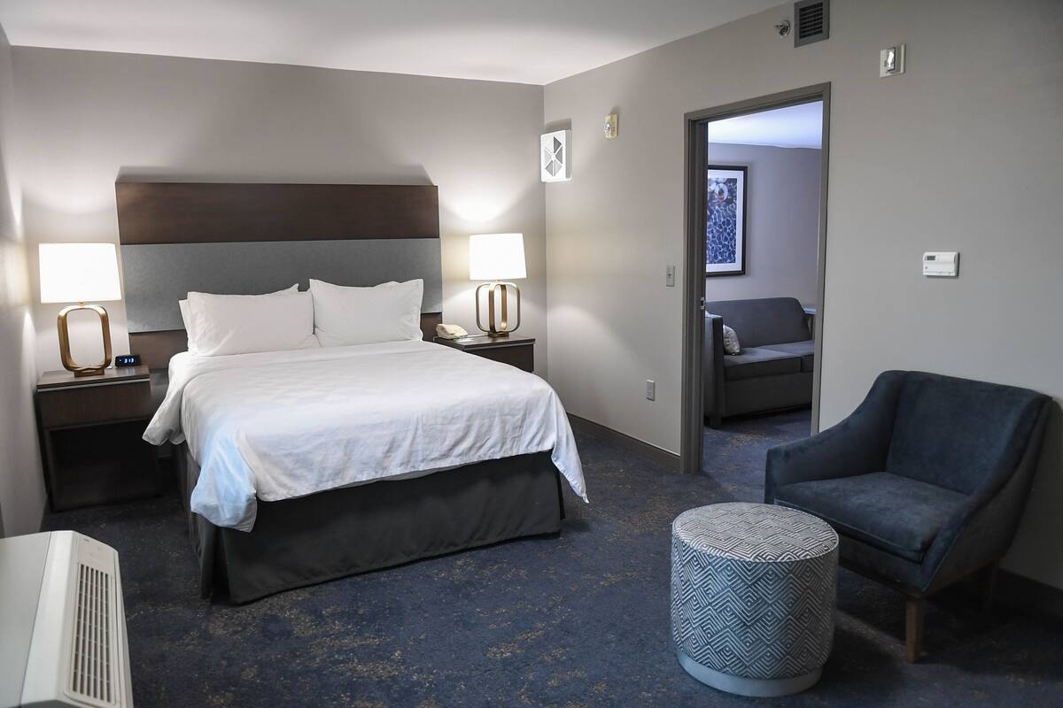 A hotel room with a large bed, two bedside tables with lamps, a chair, a round ottoman, and a door leading to another room.