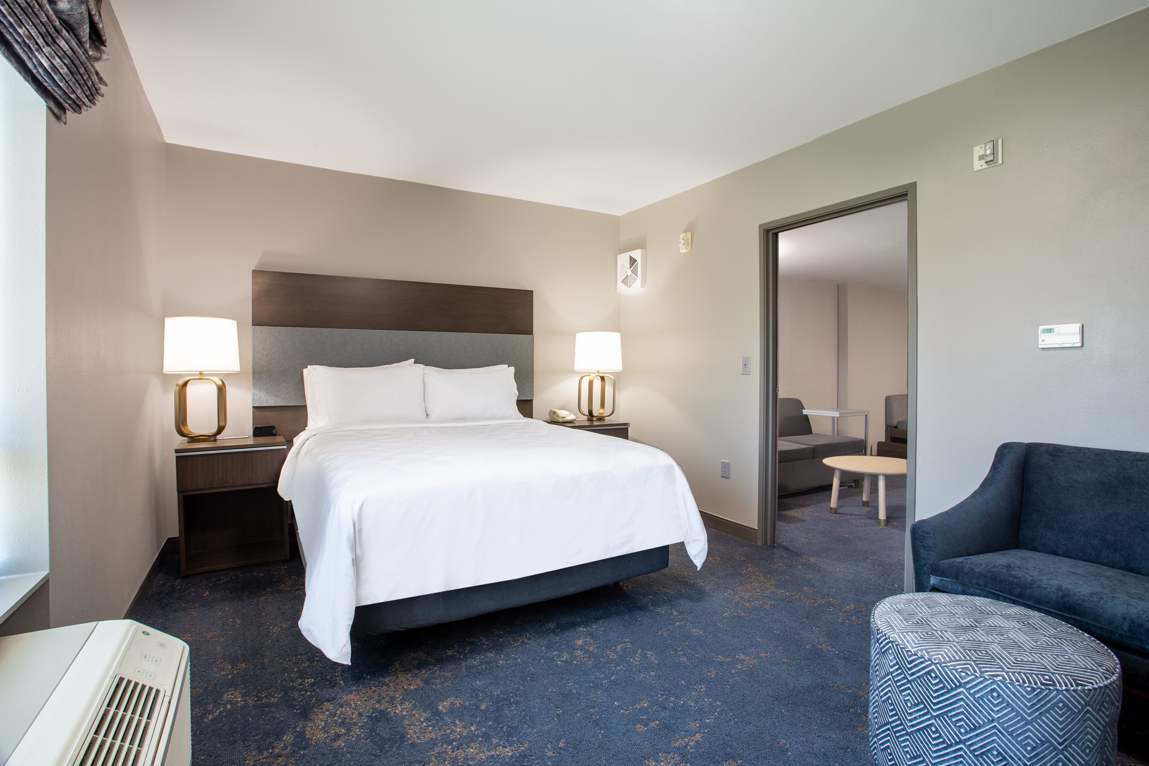 A hotel room with a large bed, two bedside lamps, a dark blue carpet, a blue sofa, and a patterned ottoman.