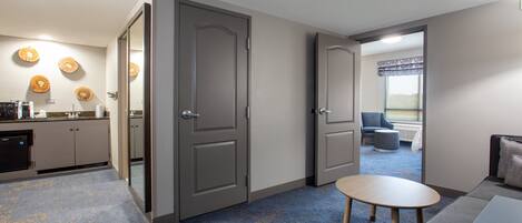 A modern hotel room with a kitchenette, a seating area with a sofa and a small table, and a view through an open door.