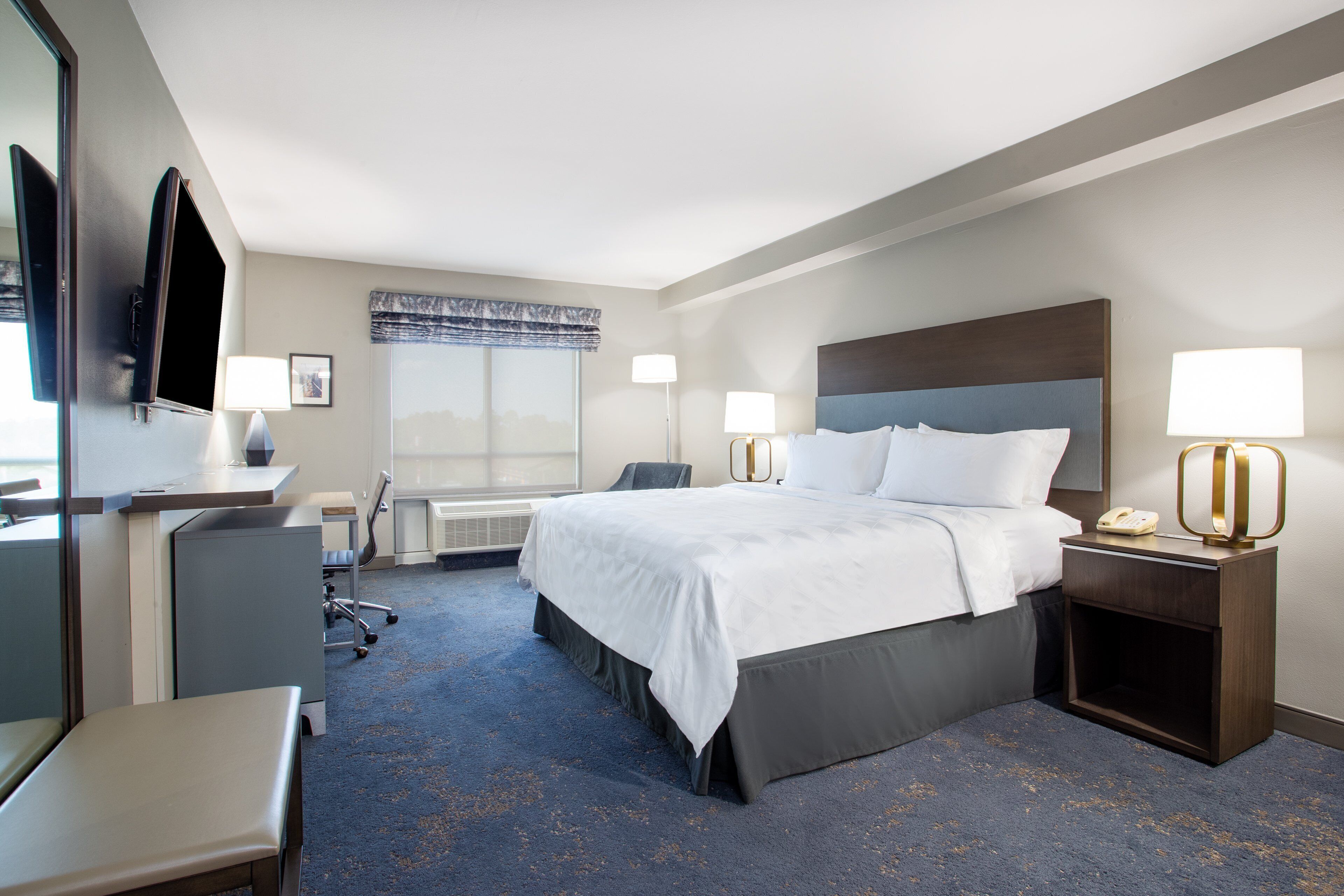 A hotel room with a large bed, two bedside tables with lamps, a desk, a chair, and a television on the wall.
