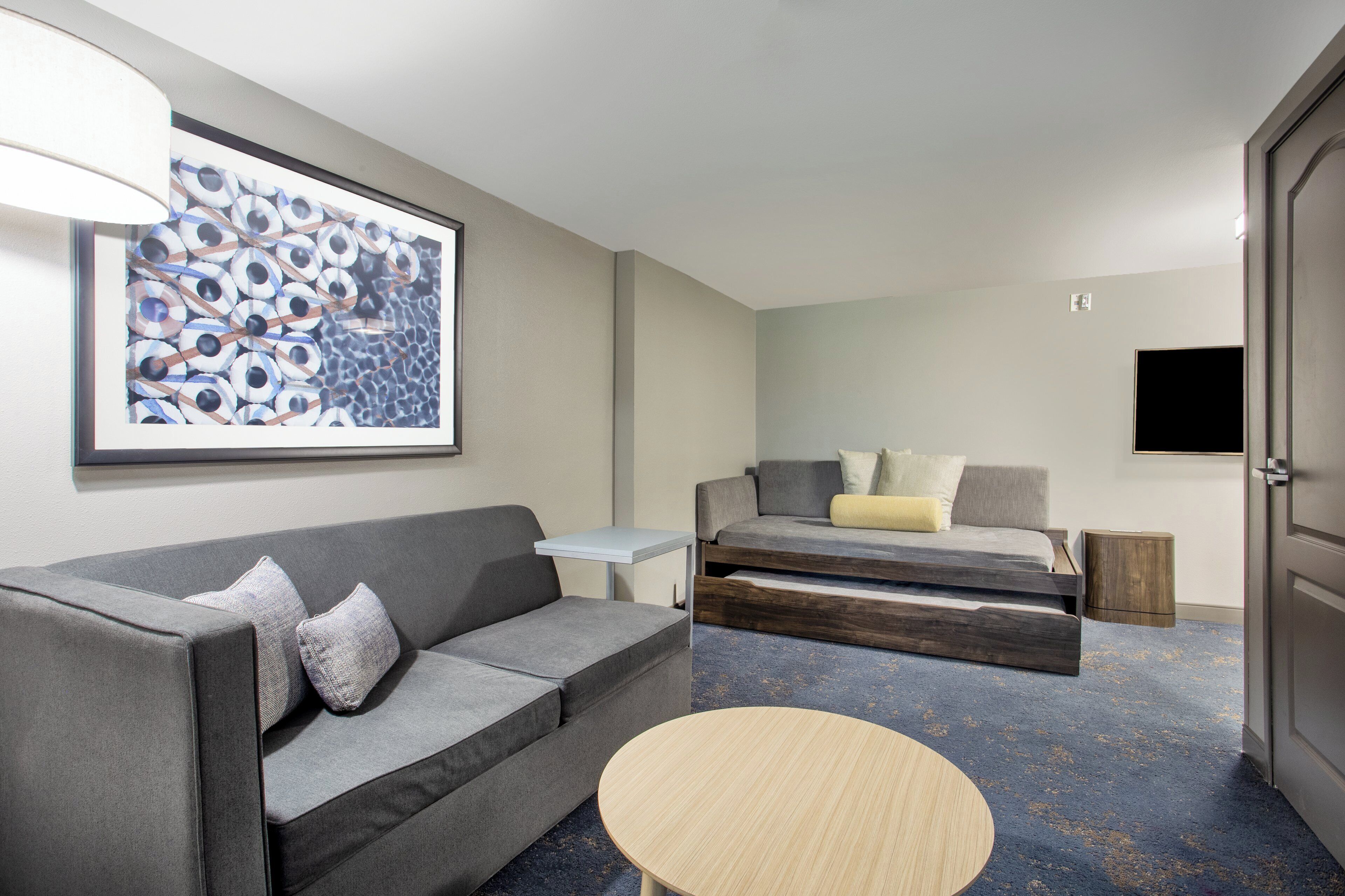 A modern hotel room with a gray sofa, a small round table, a bed with a wooden frame, and a wall-mounted TV.