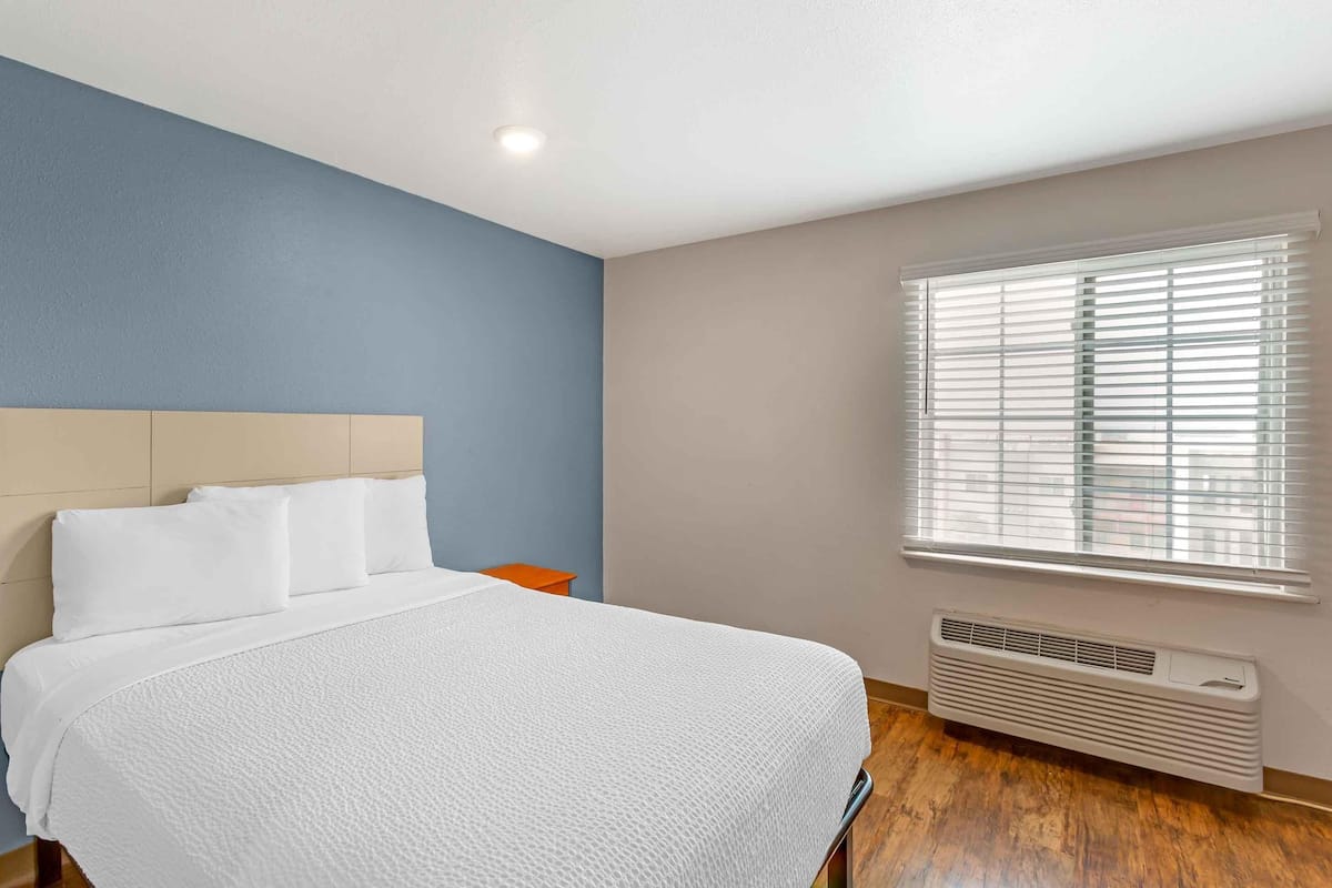 A bedroom with a bed, a nightstand, a window with blinds, and a wall-mounted heater.