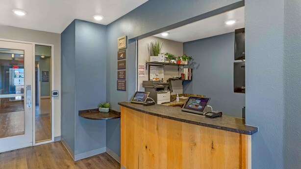 A reception desk with a wooden counter, a digital display, and a small plant. - Extended Stay America Select Suites - Las Cruces (Las Cruces)