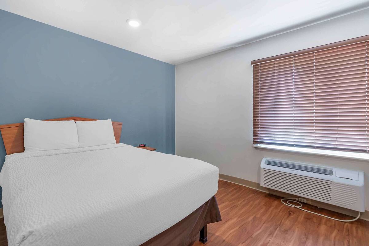 Un dormitorio con cama, mesita de noche, ventana con persianas y aire acondicionado instalado en la pared.
