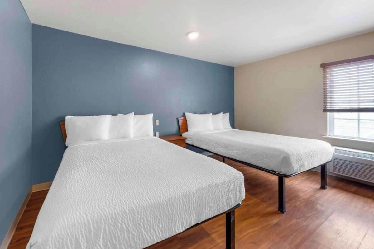 Une chambre avec deux lits, un plancher en bois, une fenêtre avec des stores et un luminaire au plafond.