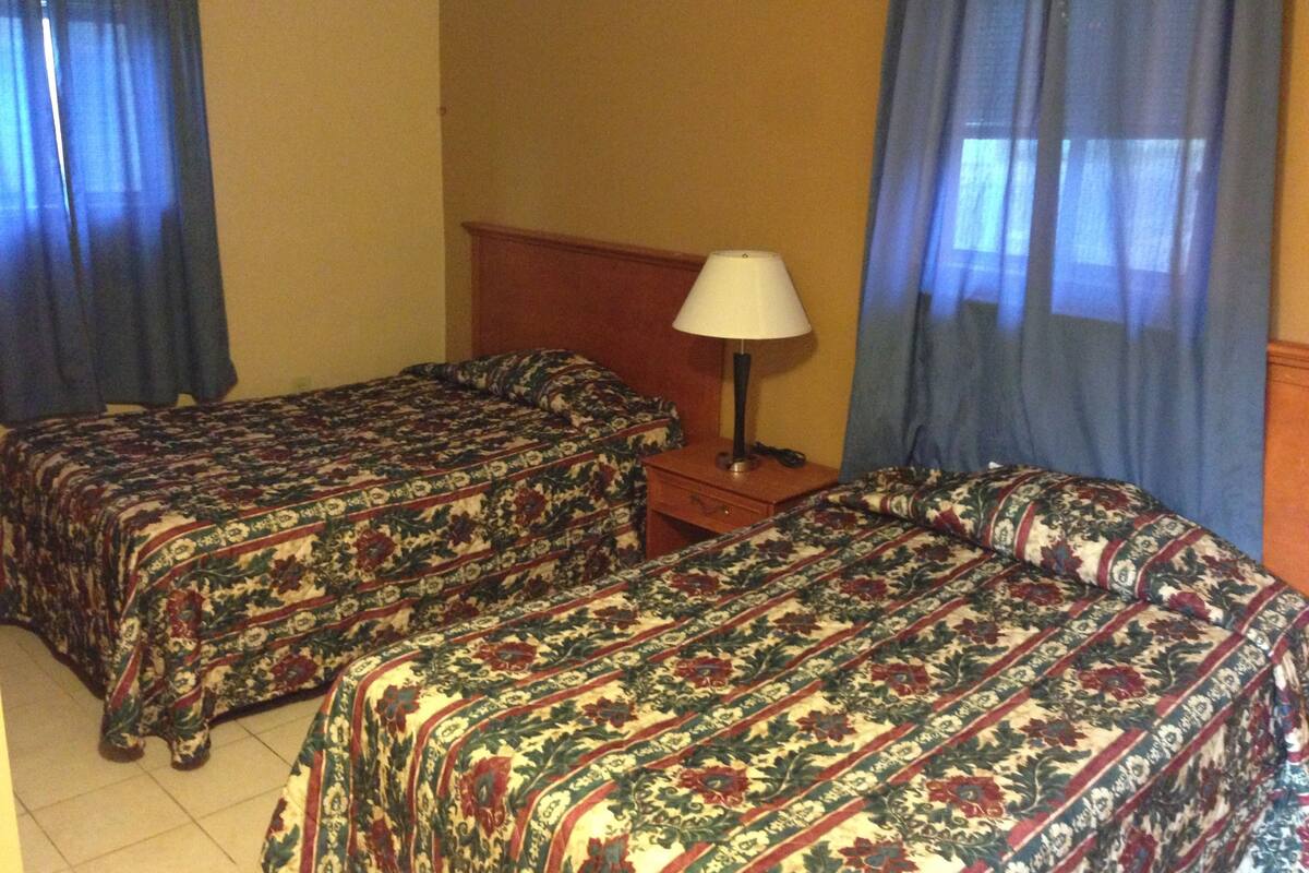 A hotel room with two single beds, a wooden nightstand, a lamp, and a window with blue curtains.