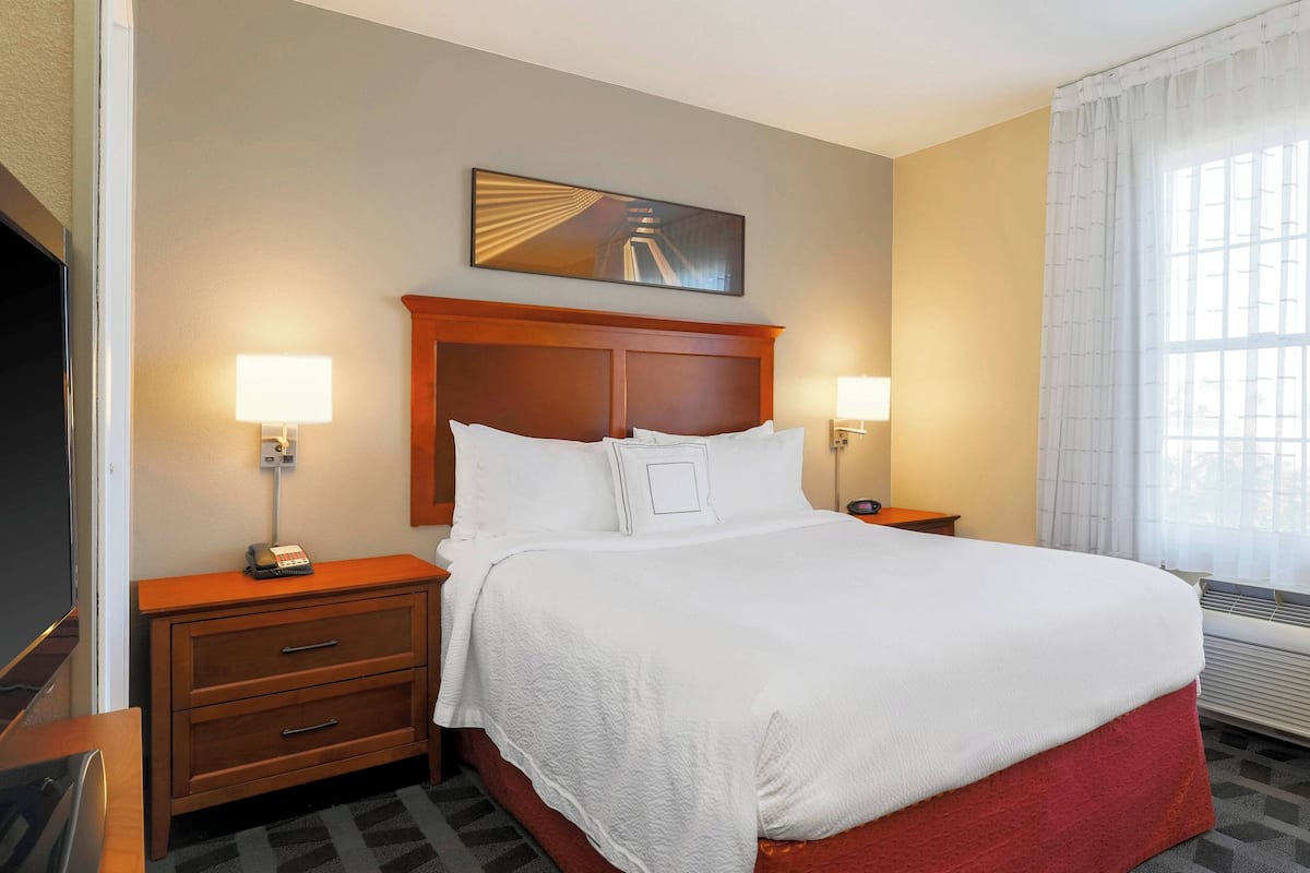 A hotel room with a large bed, two bedside tables, a flat-screen TV, a painting on the wall, and a window with curtains.