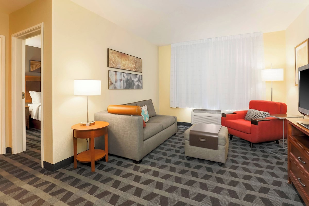 A hotel room with a sofa, a red armchair, a small table, a TV, and a window with curtains.