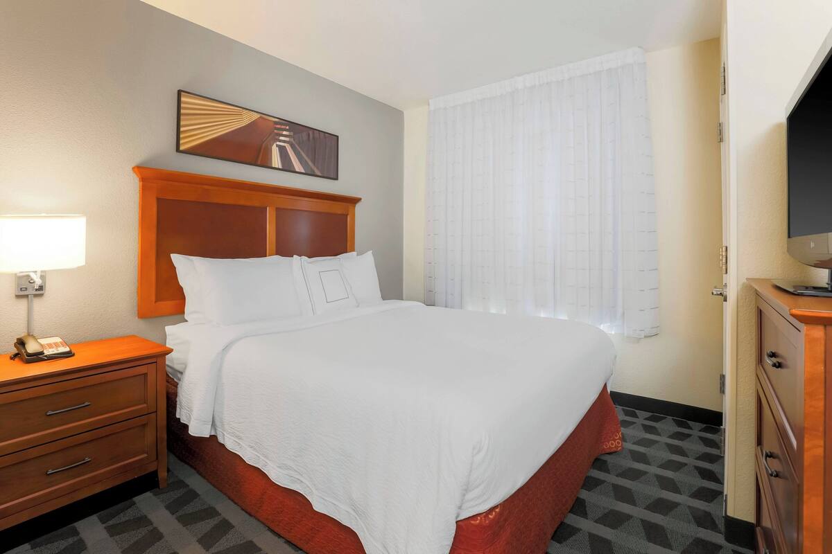 A hotel room with a bed, a wooden nightstand with a lamp, a television, and a window with curtains.
