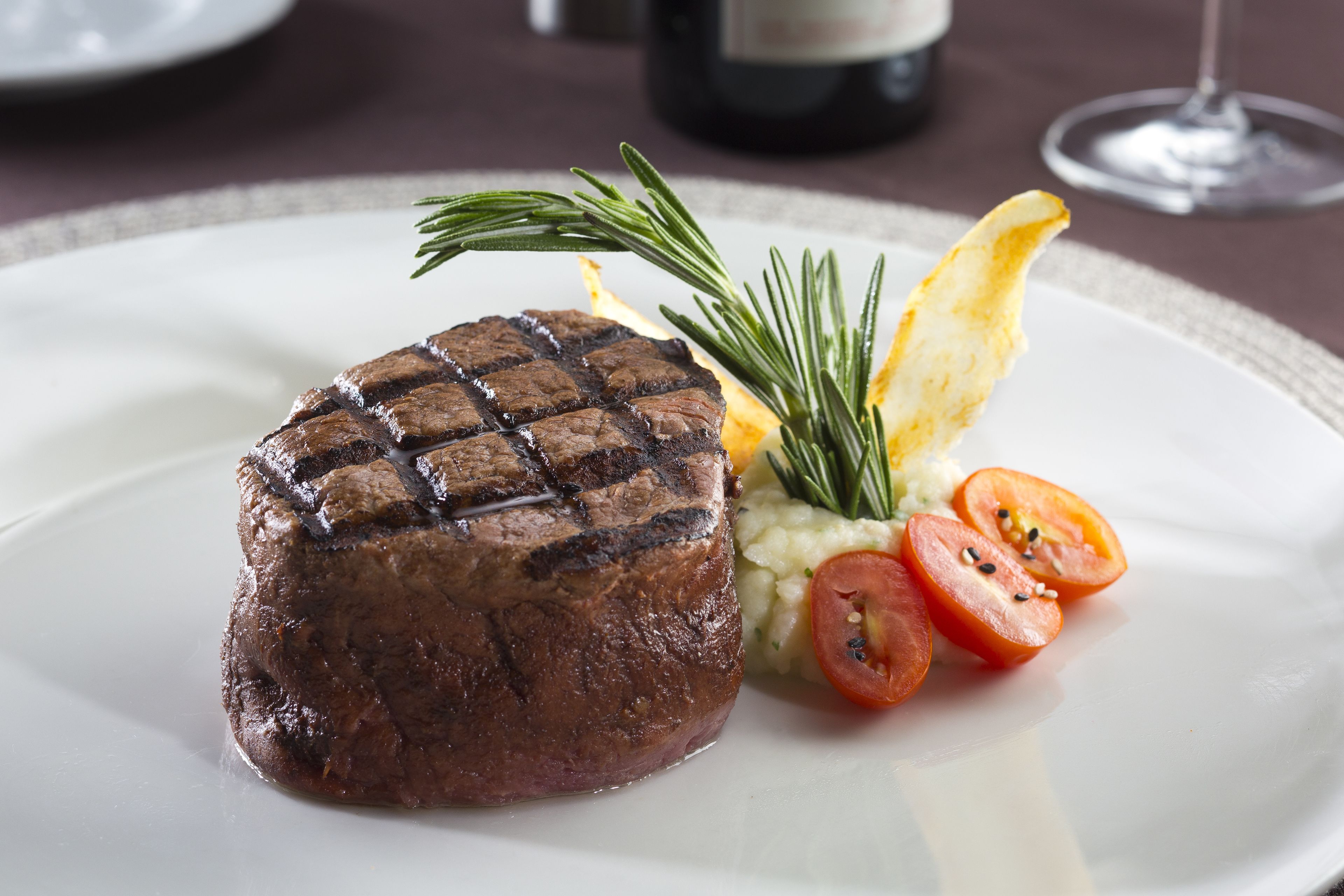 A steak with grill marks, accompanied by a sprig of rosemary and slices of tomato on a white plate.