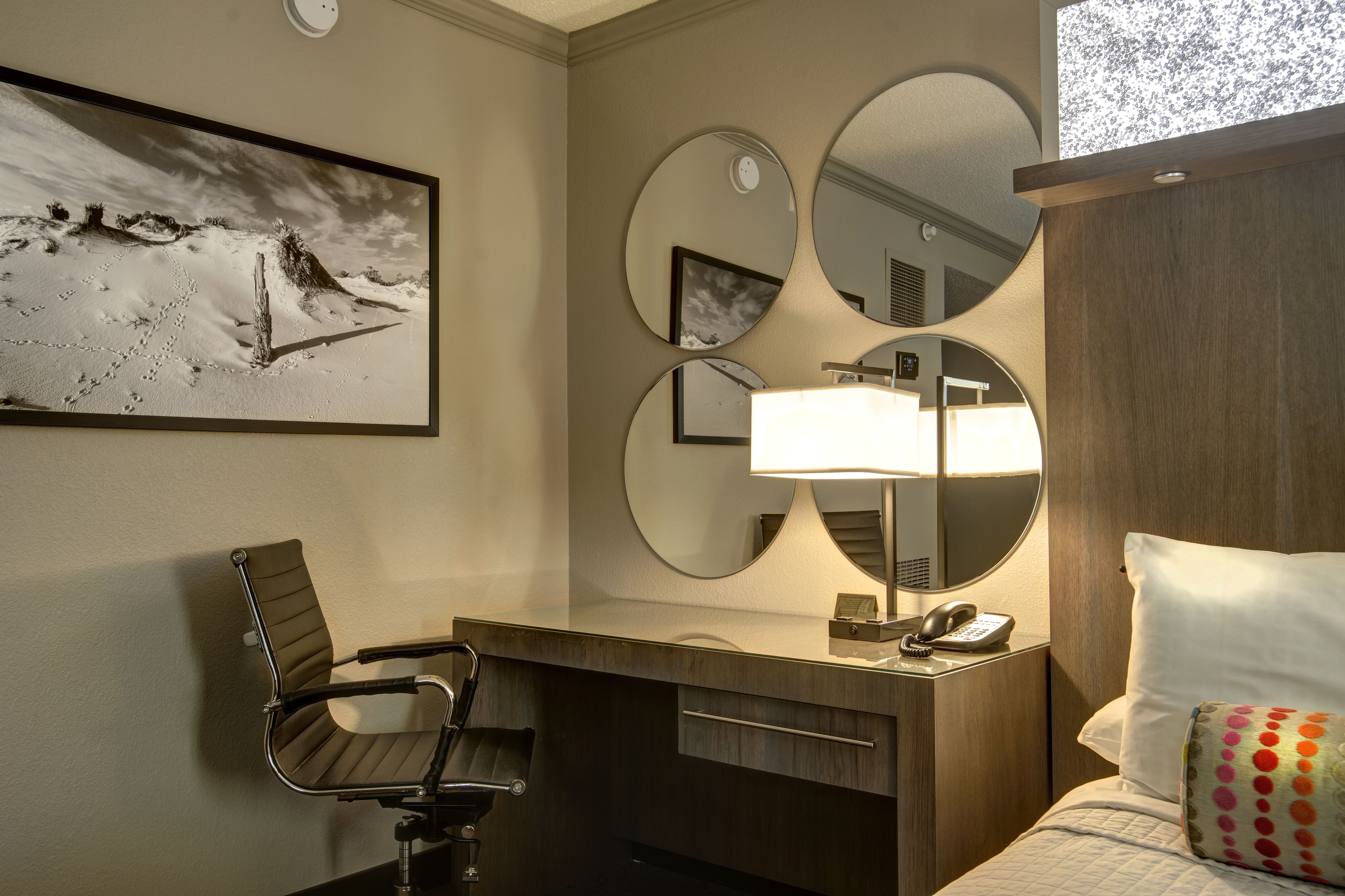 A hotel room with a bed, a desk with a chair, a desk lamp, a telephone, and three mirrors on the wall.