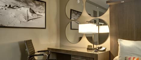 A hotel room with a bed, a desk with a chair, a desk lamp, a telephone, and three mirrors on the wall.