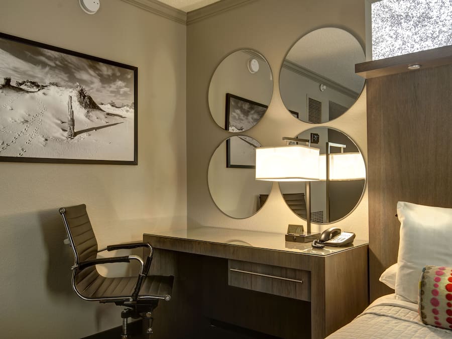 A hotel room with a bed, a desk with a chair, a desk lamp, a telephone, and three mirrors on the wall.