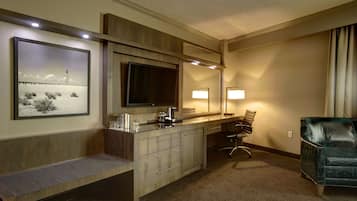 A hotel room with a wooden bench, a desk with a flat-screen TV, a chair, and a large mirror.
