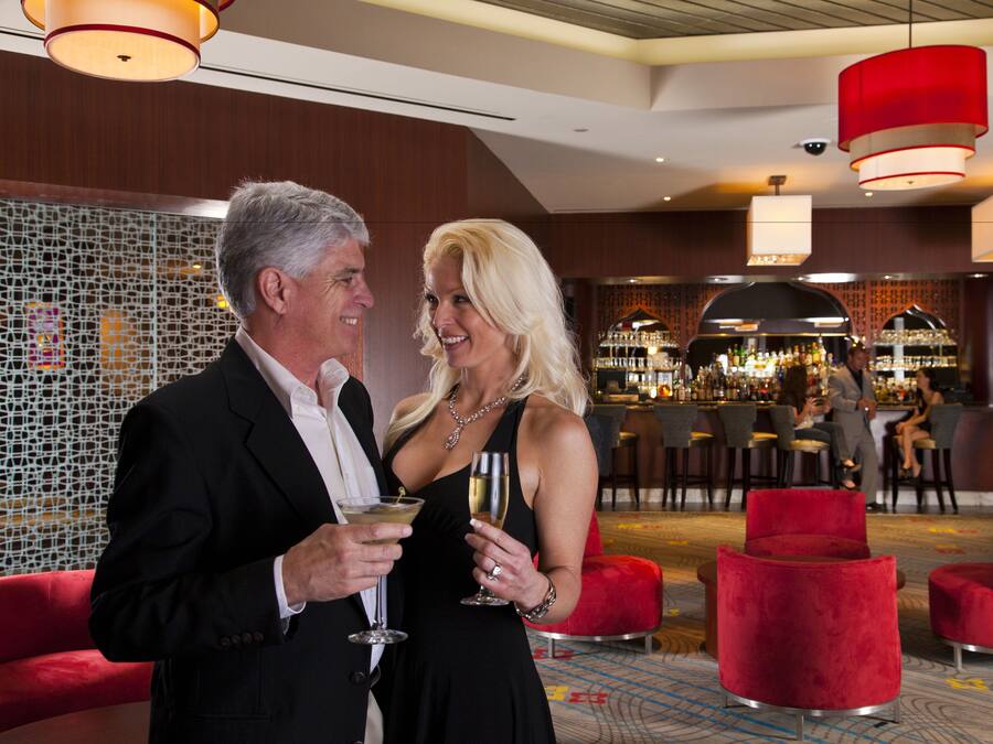 A man and woman holding drinks in a modern hotel lobby with red seating and a bar area in the background.