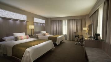 A hotel room with two beds, a desk, a chair, a TV, and a large window with curtains.