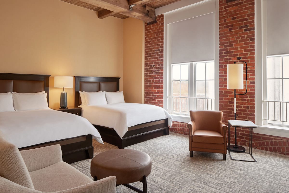 A hotel room with two beds, a chair, a table, and a lamp.