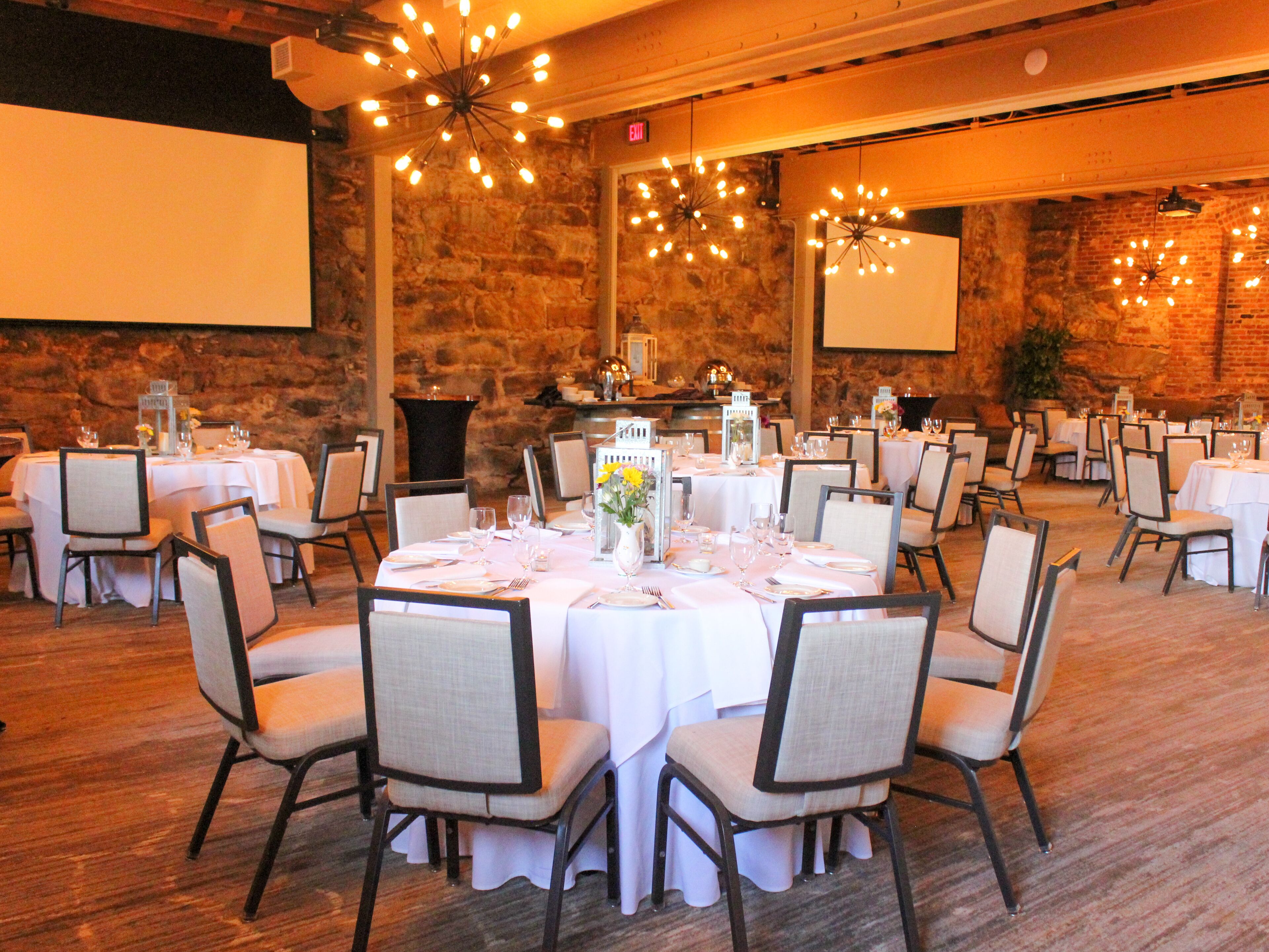 Een feestzaal met ronde tafels gedekt voor diners, stenen muren en moderne kroonluchters.