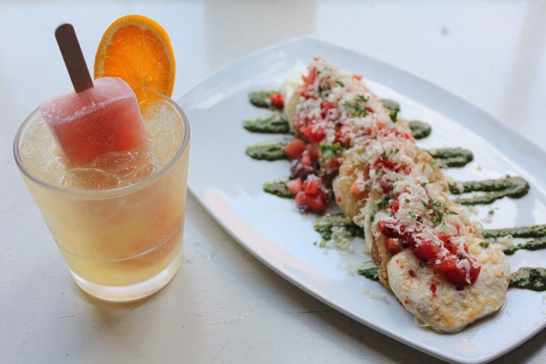 Un vaso de bebida helada con una rodaja de fruta y una pajita, y un plato con comida decorada con hierbas y tomates.