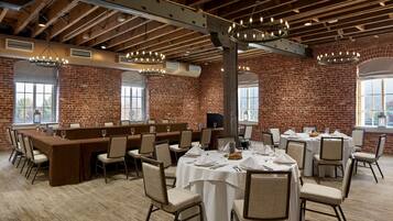 A spacious dining area with high ceilings, exposed beams, and brick walls. Tables are set with white tablecloths and chairs, and there are large windows providing natural light.