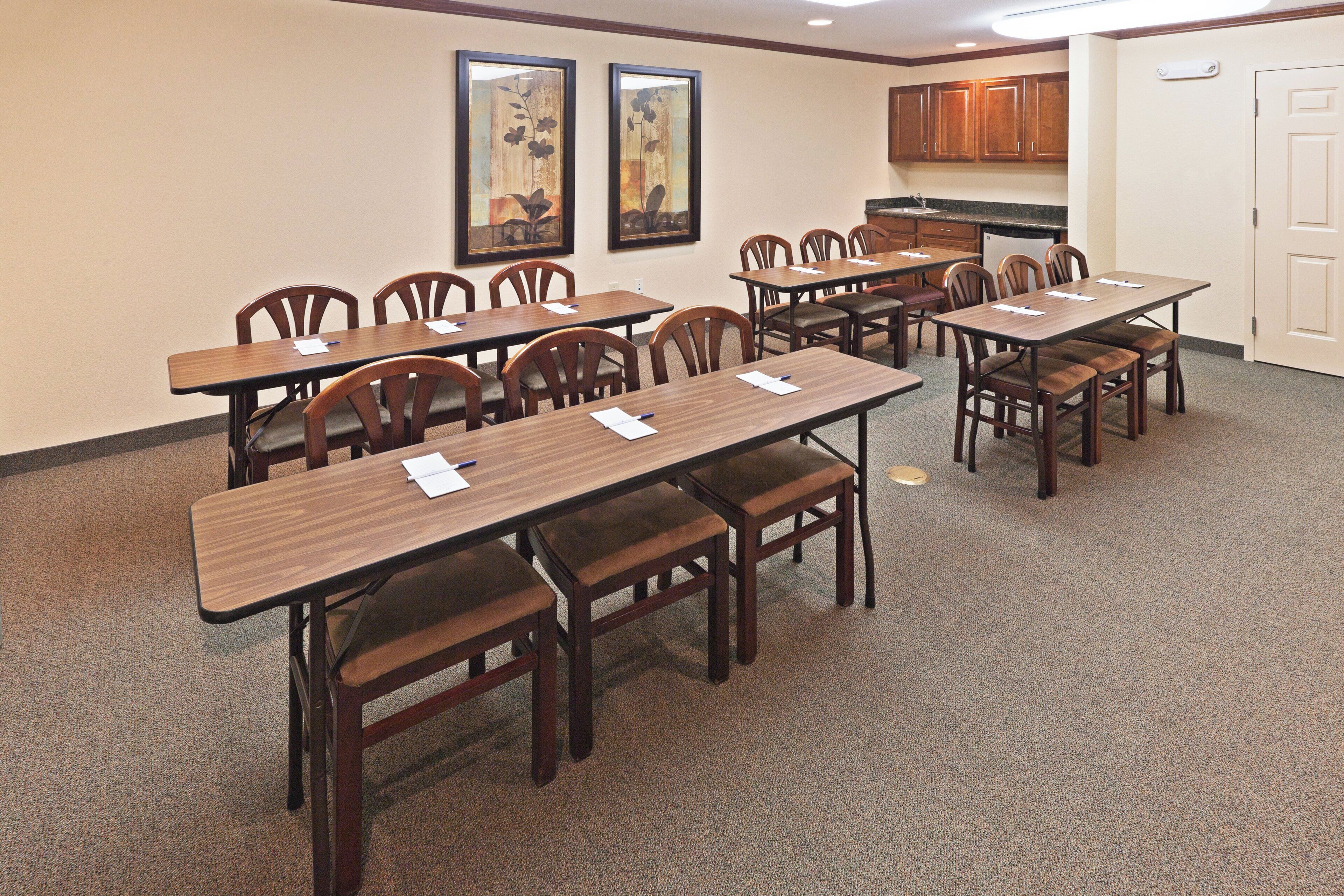 A conference room with multiple tables and chairs, a kitchenette, and two framed artworks on the wall.