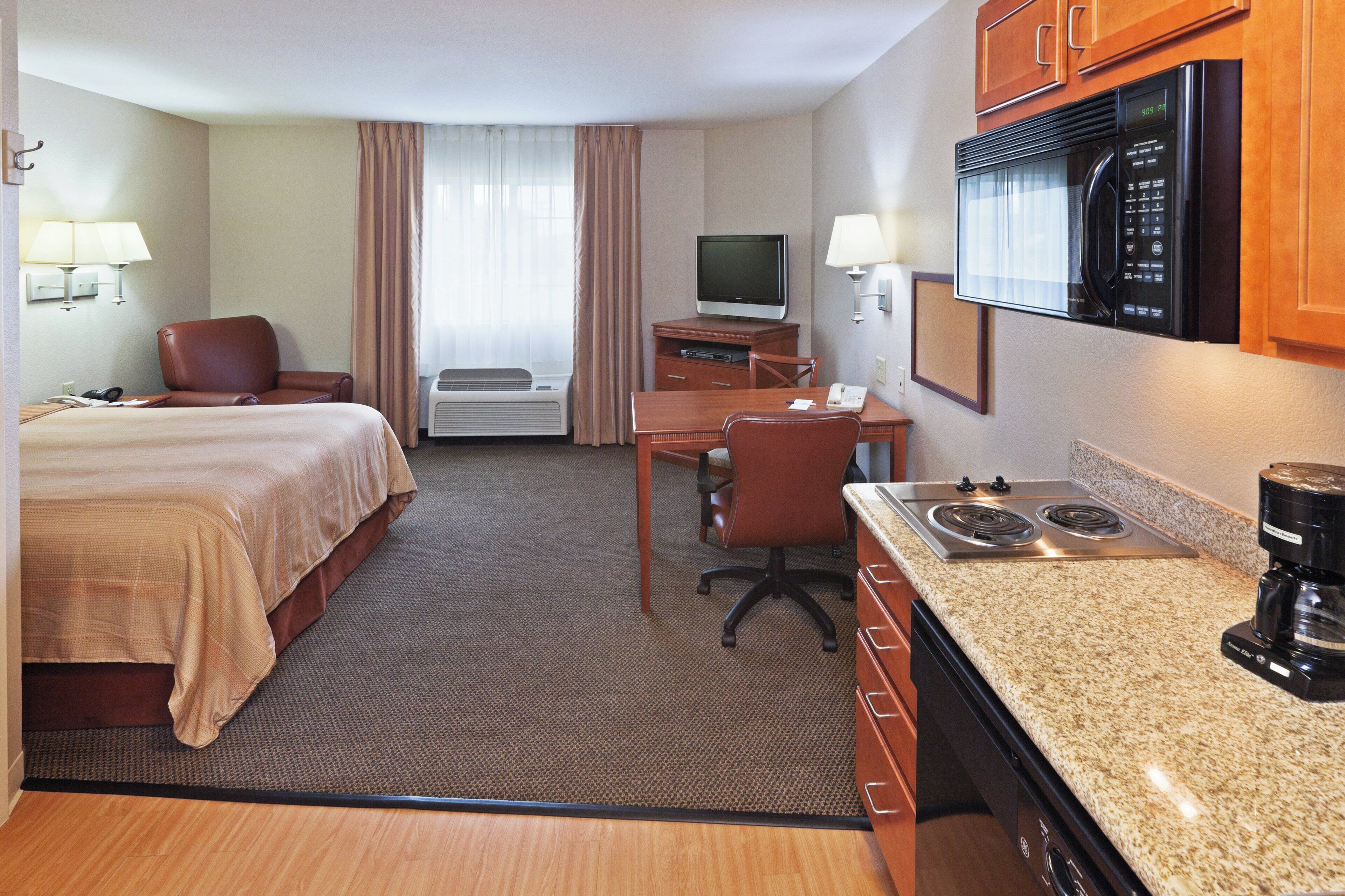 A hotel room with a bed, desk, chair, kitchenette, television, and air conditioning unit.