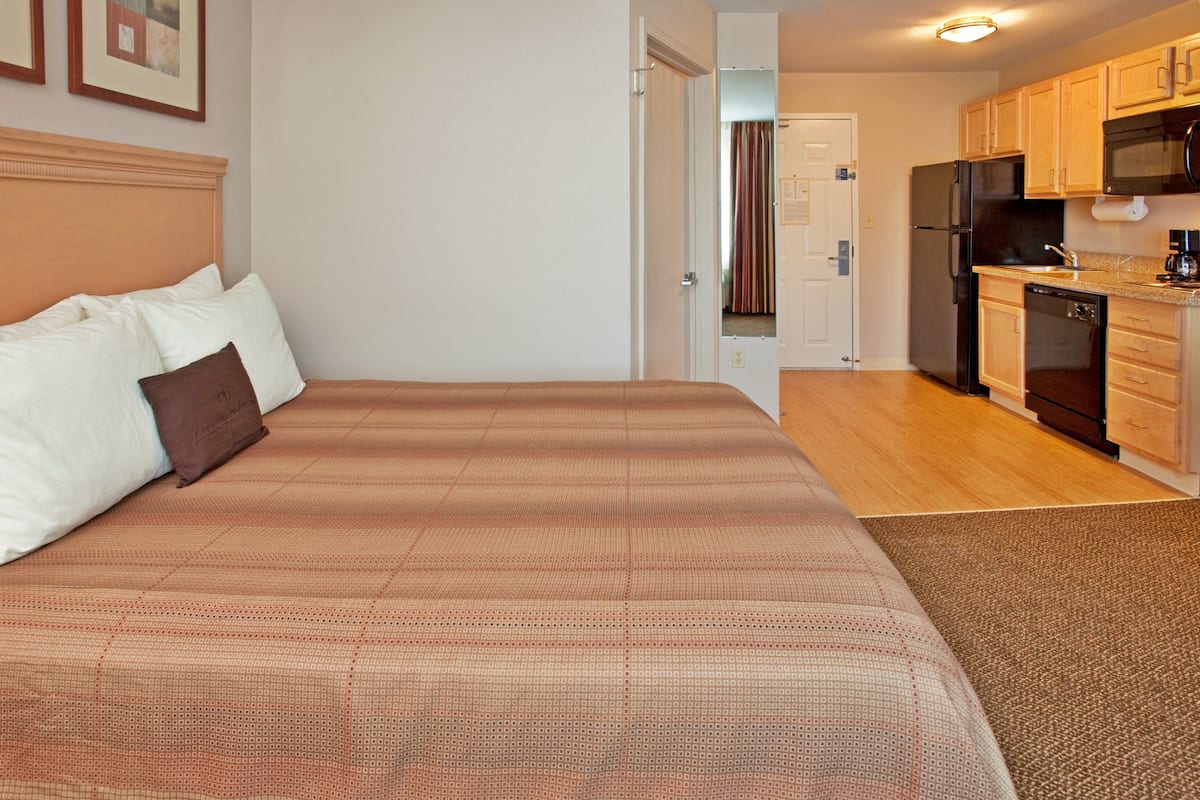Una habitación de hotel con una cama grande, cabecera de madera, y una cocina equipada con refrigerador, microondas y lavavajillas.