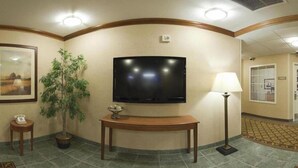 Eine Hotellobby mit einem an der Wand befestigten Flachbildfernseher, einem Holzkonsolentisch, einem Topfpflanzen, einer Stehlampe und einem gerahmten Bild.