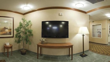 Un vestíbulo de hotel con un televisor de pantalla plana montado en la pared, una mesa consola de madera, una planta en maceta, una lámpara de pie y un cuadro enmarcado.