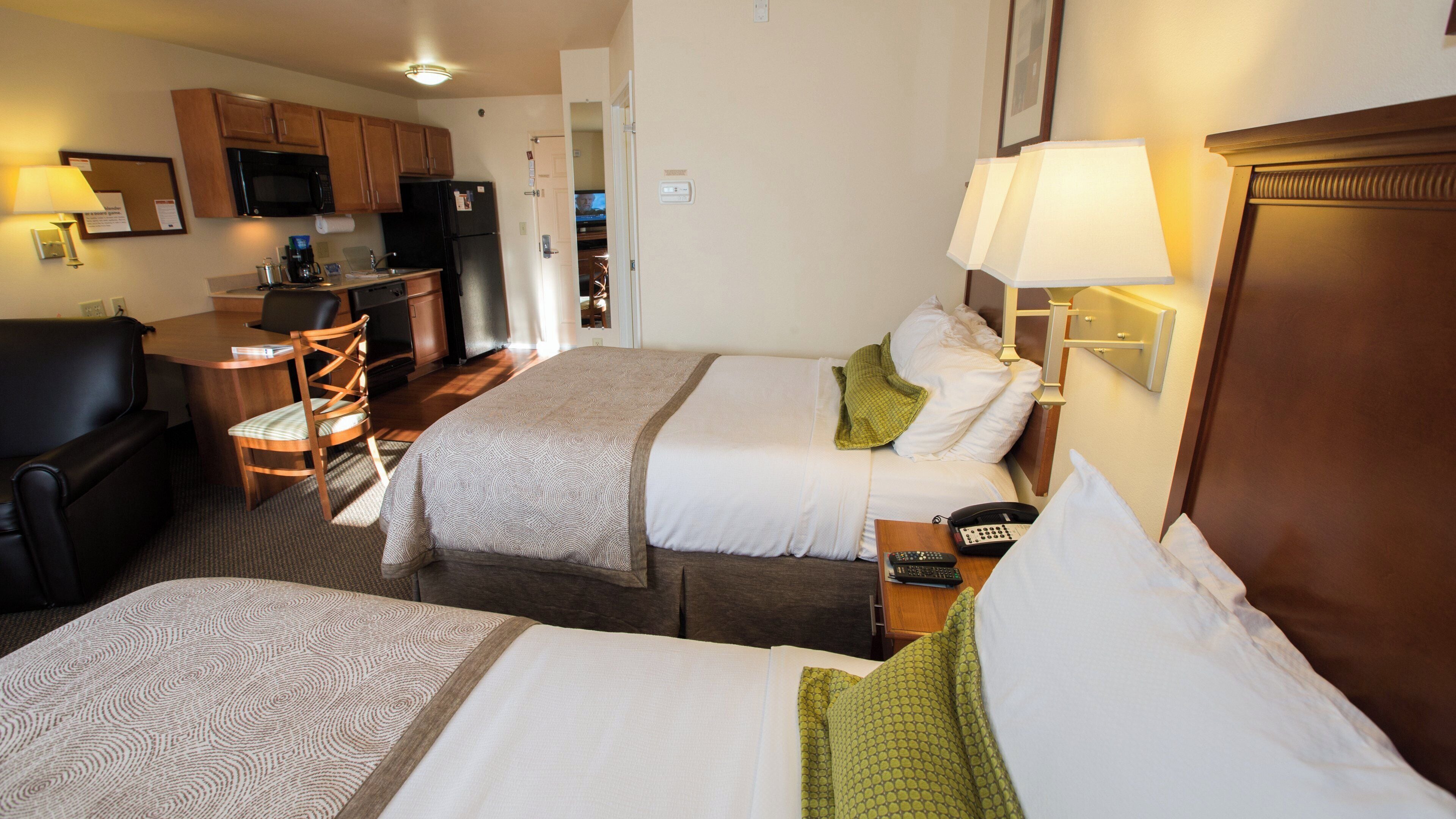 A hotel room with two beds, a desk, a chair, a microwave, and a small kitchenette.