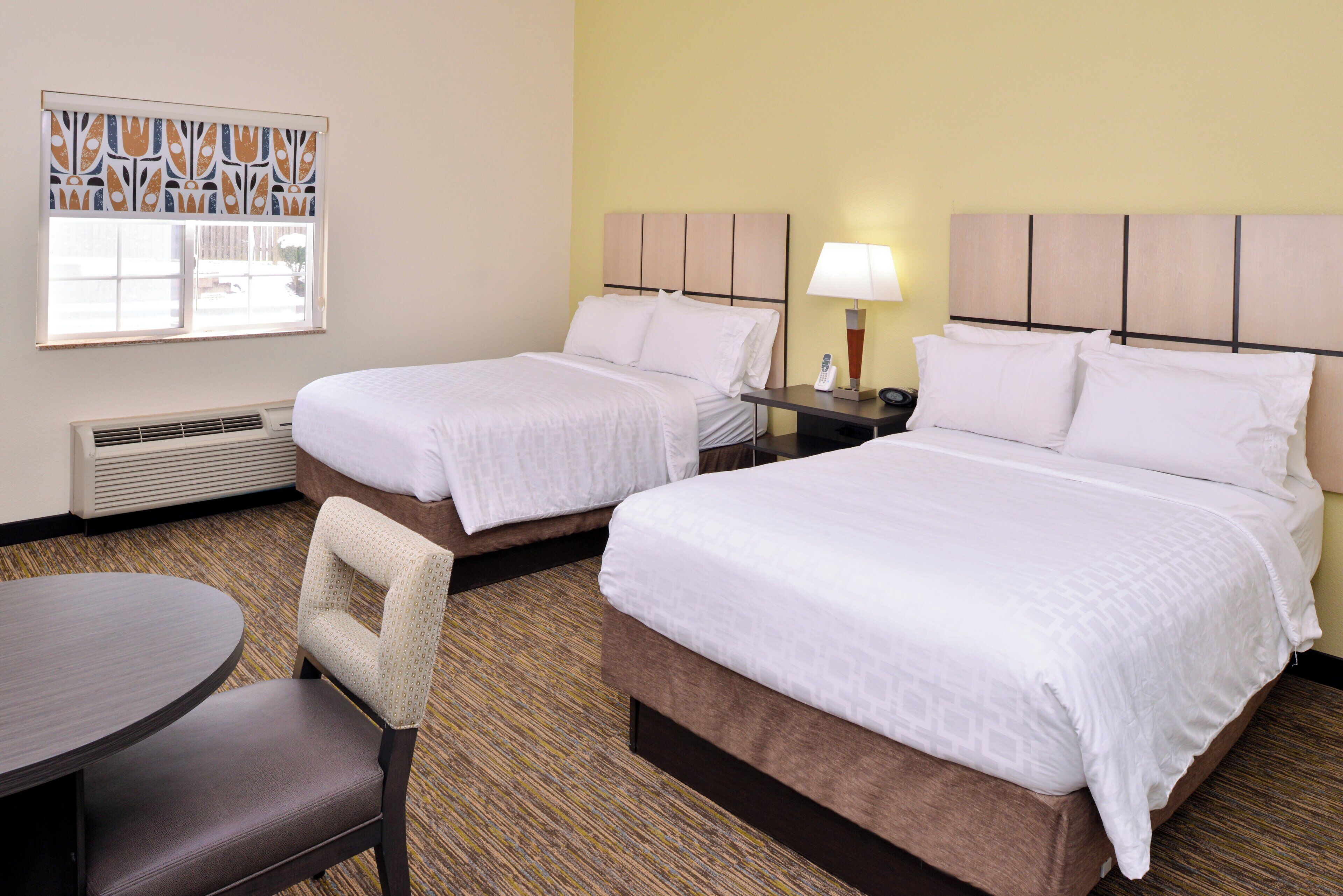 A hotel room with two beds, a table, a chair, a window with blinds, and a lamp on the nightstand.