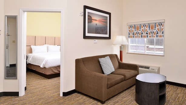 Habitación de hotel con una cama, un sofá marrón, una mesa redonda, una lámpara, un ventanal con celosía decorativa y un cuadro en la pared.