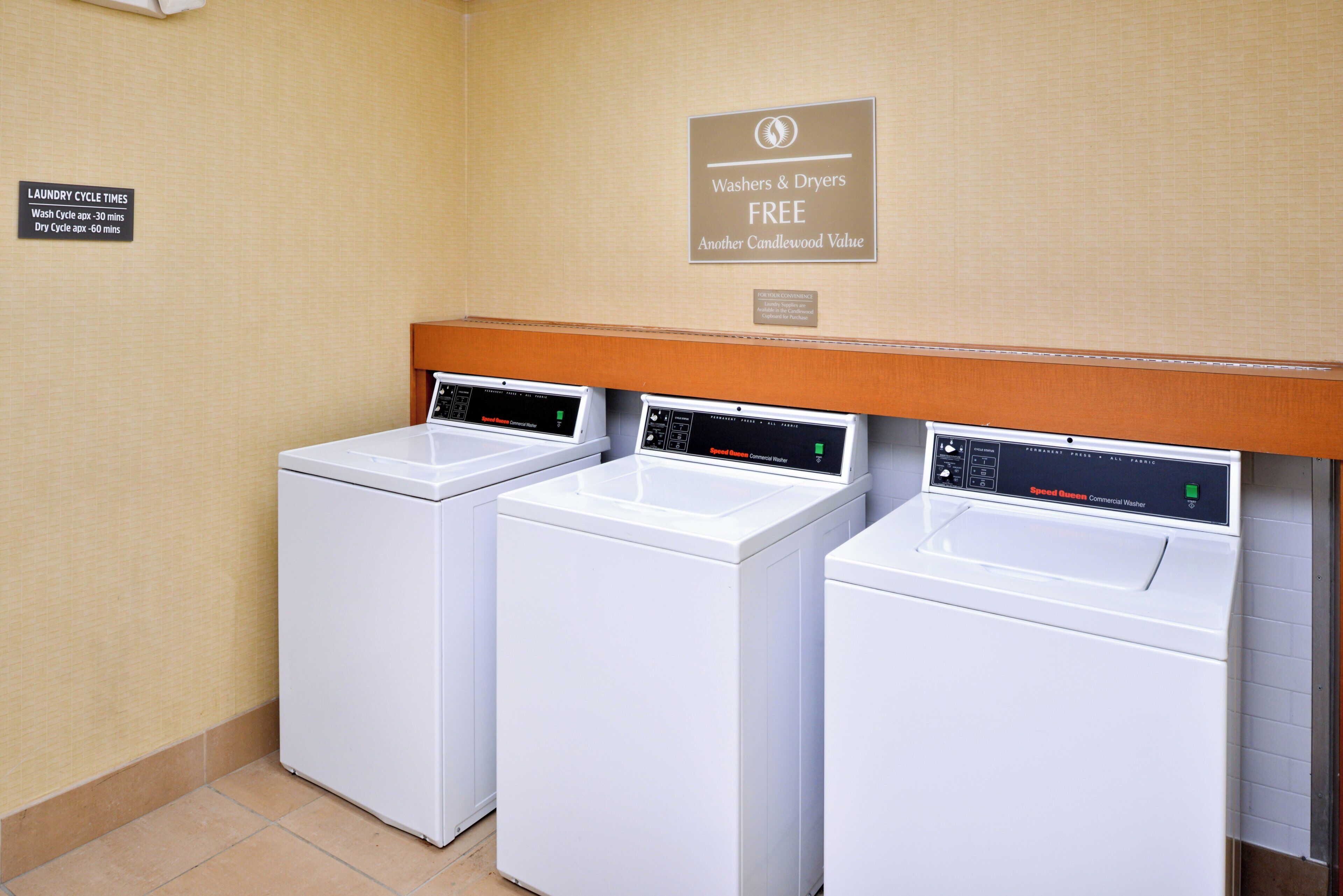 A row of three Speed Queen commercial washers in a laundromat with a sign above stating 'Washers & Dryers FREE Another Candlewood Value' and a notice about laundry supplies available for purchase.
