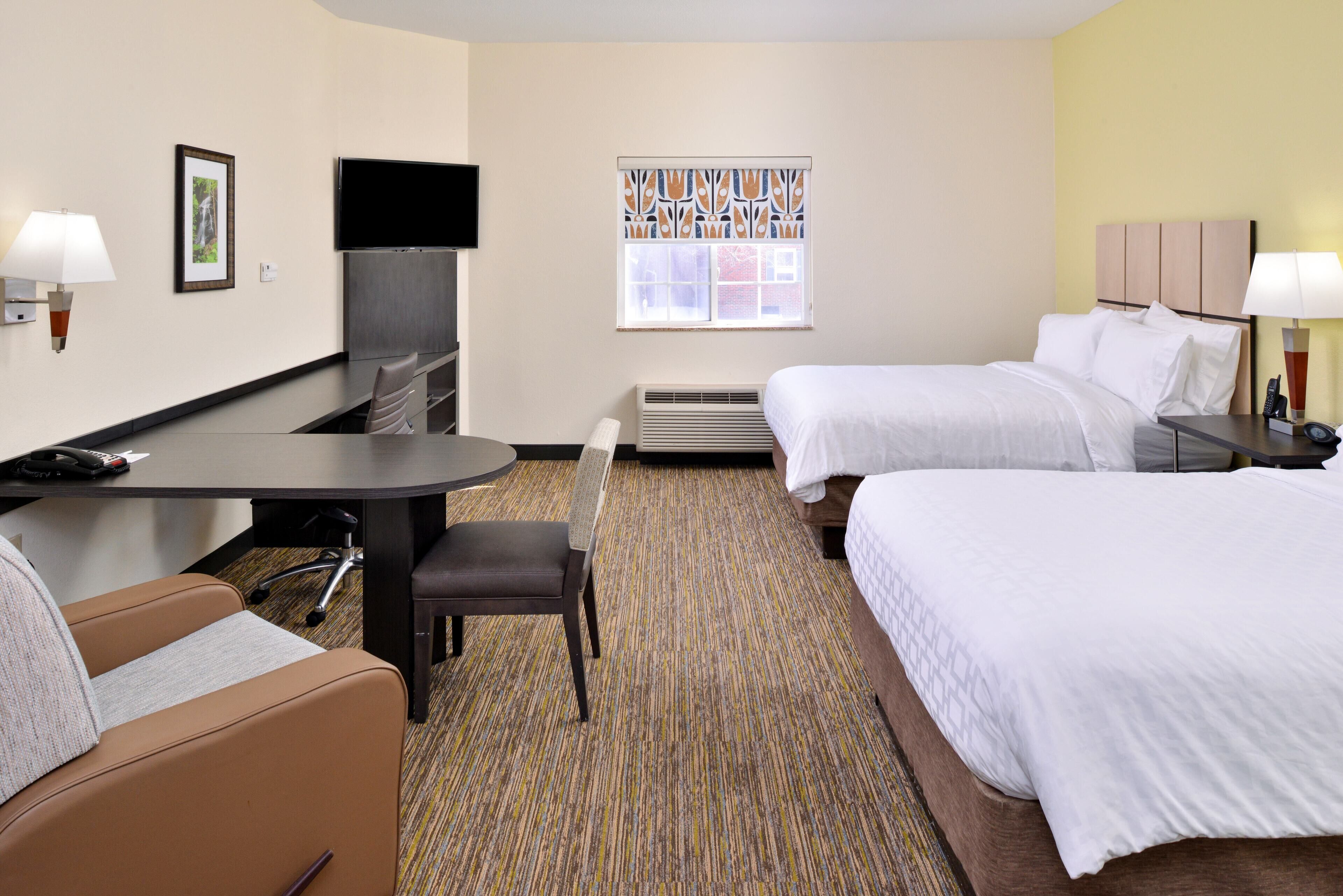 A hotel room with a desk, two beds, a TV, a chair, and a window with a patterned curtain.