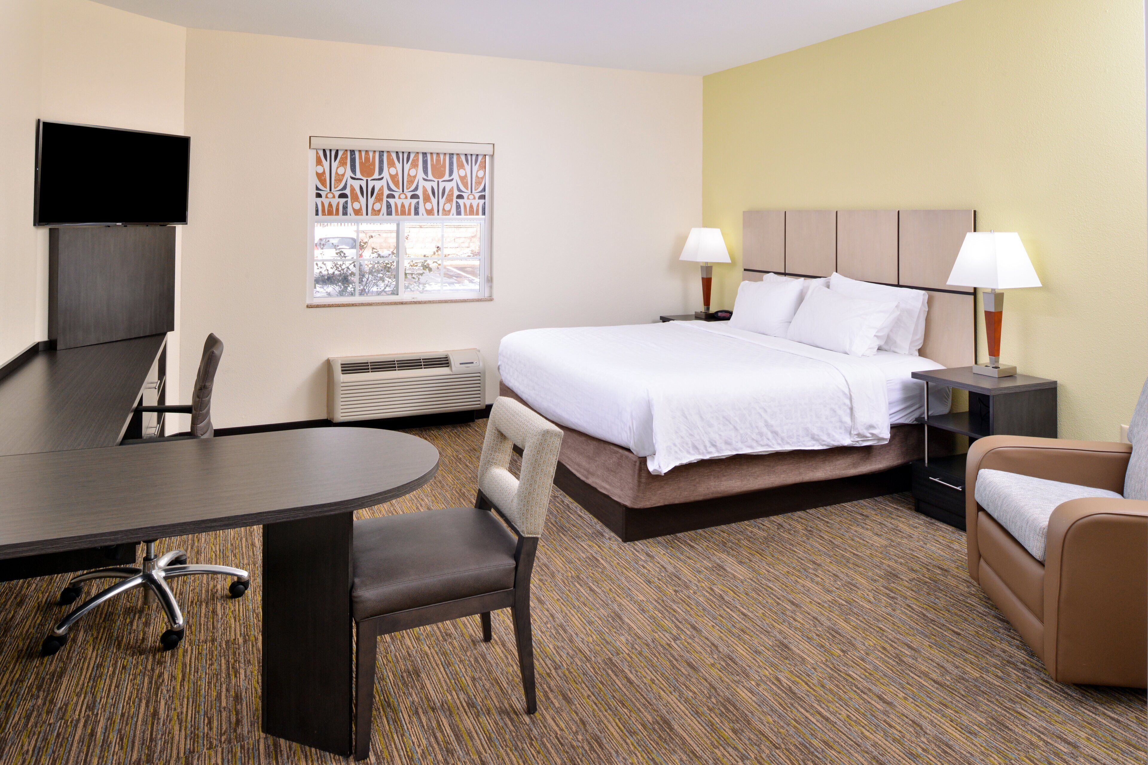 A hotel room with a bed, two bedside tables with lamps, a desk with a chair, a TV mounted on the wall, and a window with a view.
